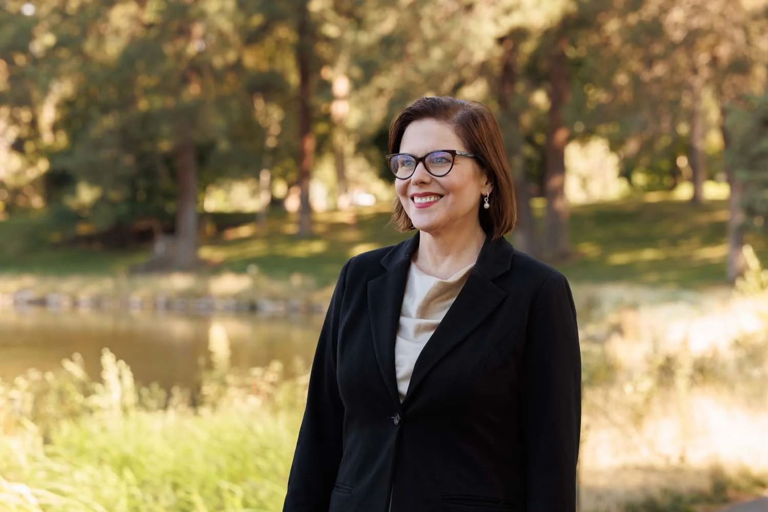Sonia Huntsman smiling outdoors near a river with trees and autumn foliage in the background.
