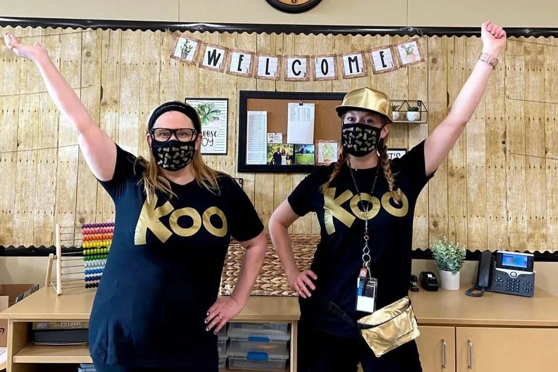 Sonia Hunstsman wearing black shirts with gold letters, matching face masks, and one with a gold hat, standing in an office or classroom with a 'Welcome' banner and celebrating.