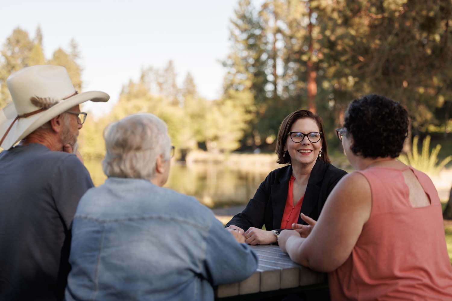 Sonia and adults sitting at a table outdoors, engaged in conversation, with trees and a pond in the background.