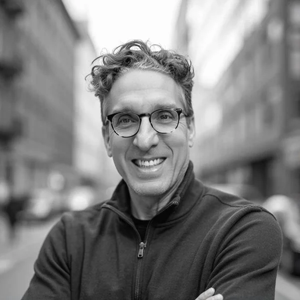 A man smiling and wearing glasses standing in a city.