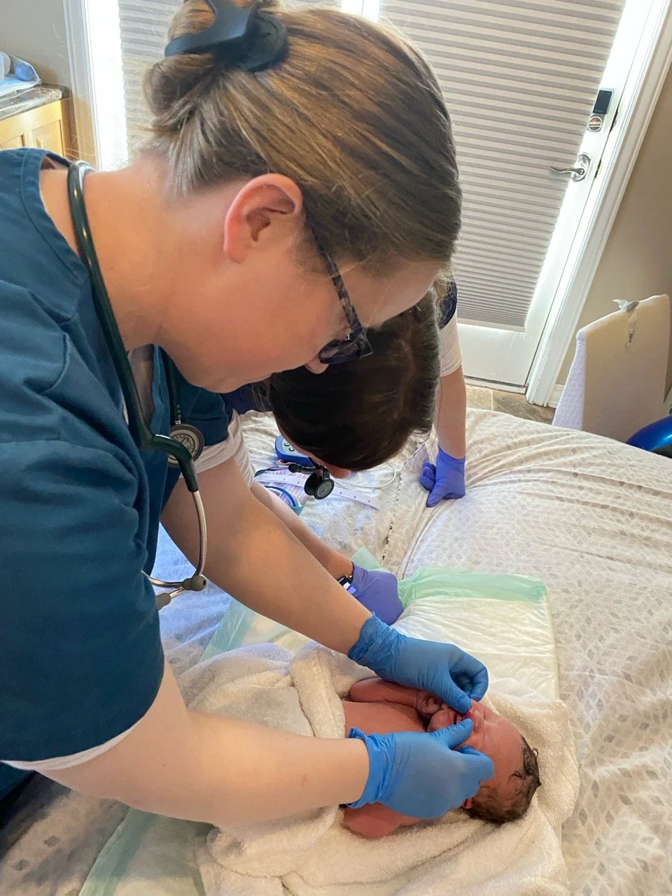 Healthcare professionals, including a nurse with glasses and a stethoscope, attending to a newborn baby on a bed, with the baby's head being carefully examined or supported.