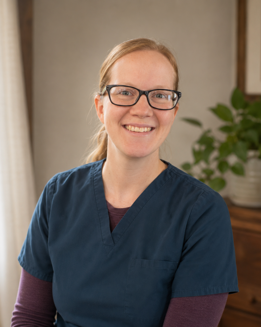 A smiling woman with glasses in medical scrubs, seated indoors, with a plant and wooden furniture in the background.