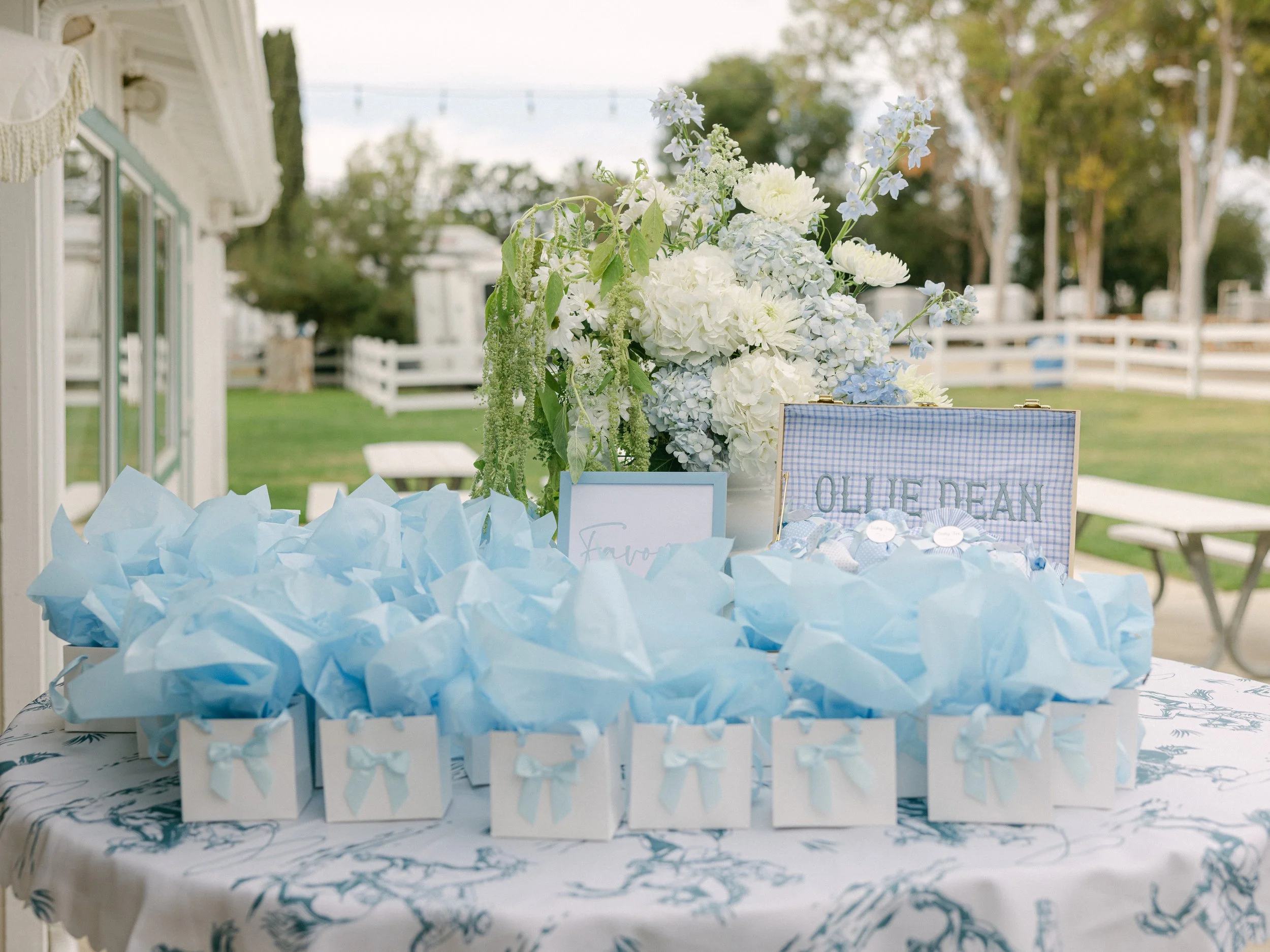 A table with several white gift boxes decorated with pale blue ribbons and tissue paper, a large floral arrangement of white and light blue flowers, and a small sign that reads 'QUINN' in front of a house with a white fence and green outdoors