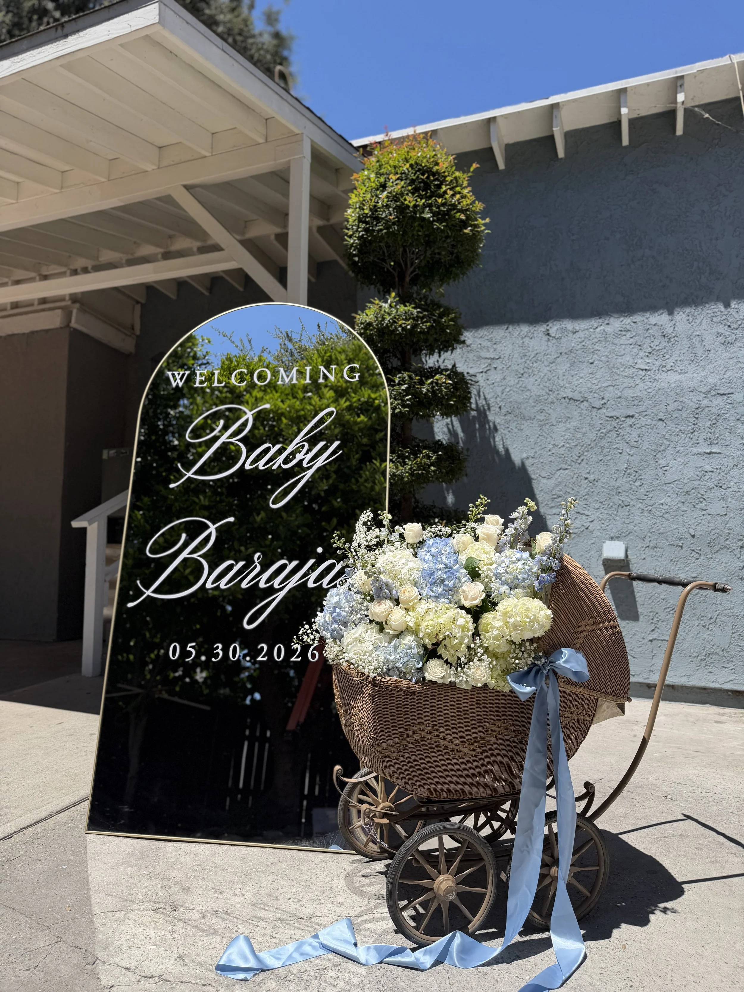 A decorative sign with the words "Welcoming Baby Brayden" and a date of 05.30.2026, positioned outdoors next to a vintage-style stroller filled with a large bouquet of white and pale blue flowers, with a light blue ribbon tied around it, against a textured light gray wall and partly cloudy blue sky.