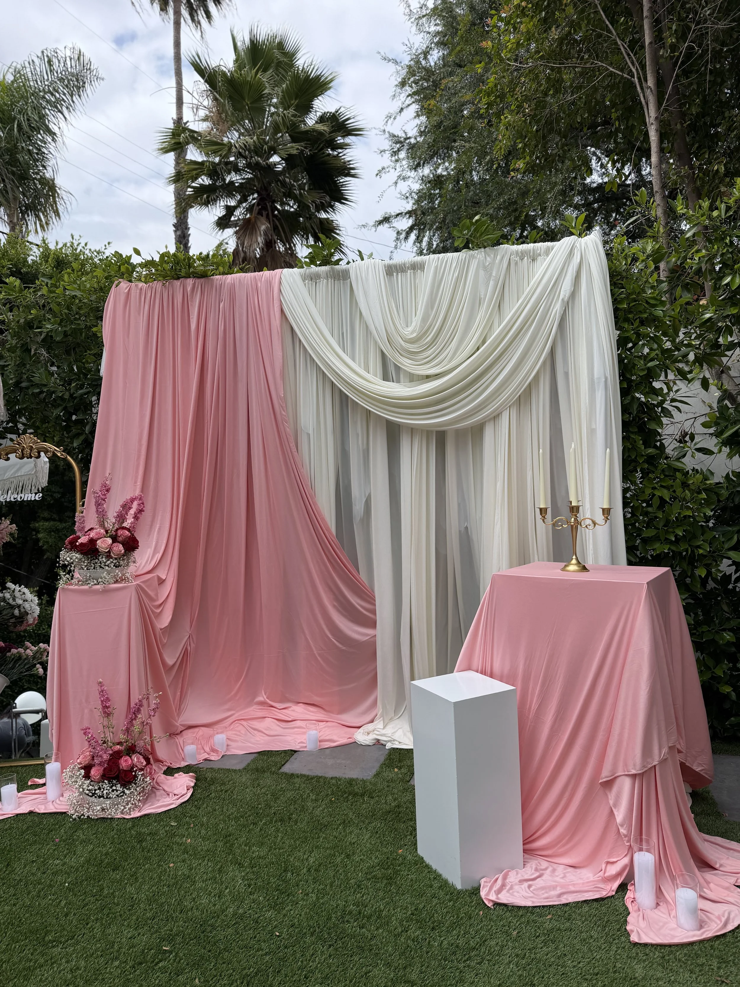 Outdoor wedding setup with pink and white draped fabrics, floral arrangements, candles, and a candelabrum on a pink-covered table, surrounded by greenery.