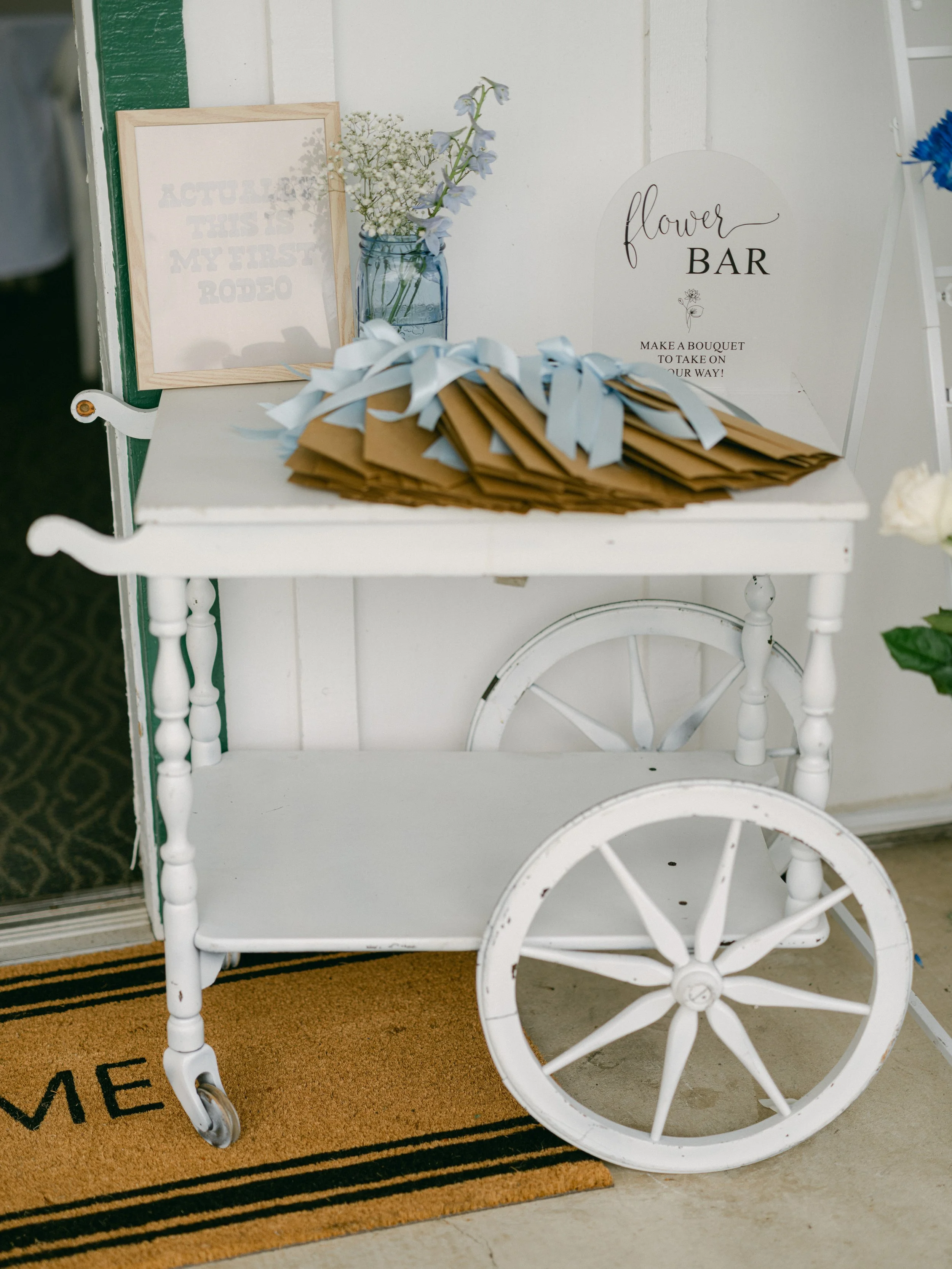 A white wooden cart with two wheels, holding a vase with white and purple flowers, and a sign that reads 'flower BAR'. There are also paper bags with blue ribbons placed on the cart, and a framed sign and a rug with a brown border on the floor.