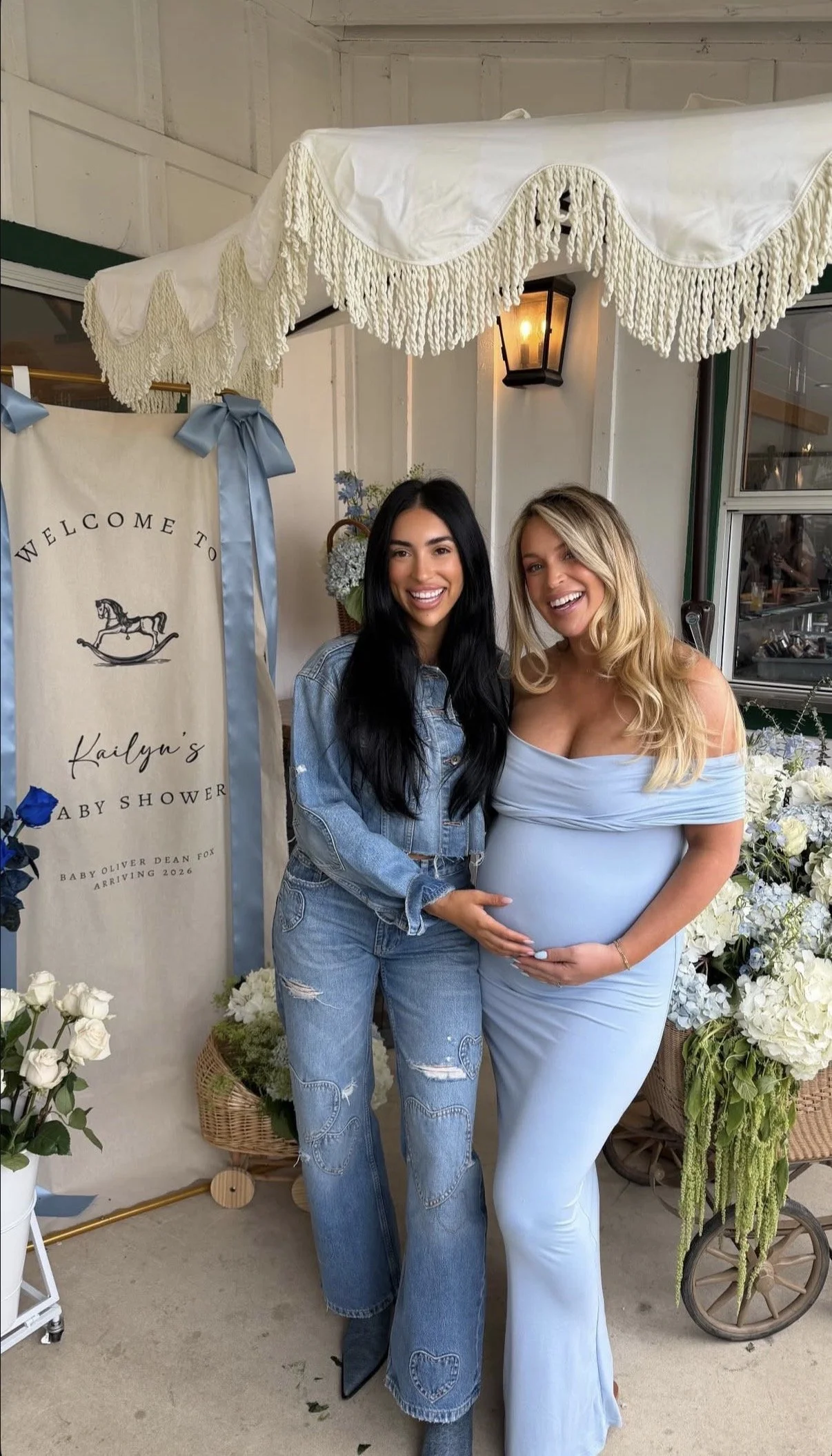 Two women standing together at a baby shower, one pregnant, in front of floral decorations and a sign that reads "Welcome to Kailyn's Baby Shower."