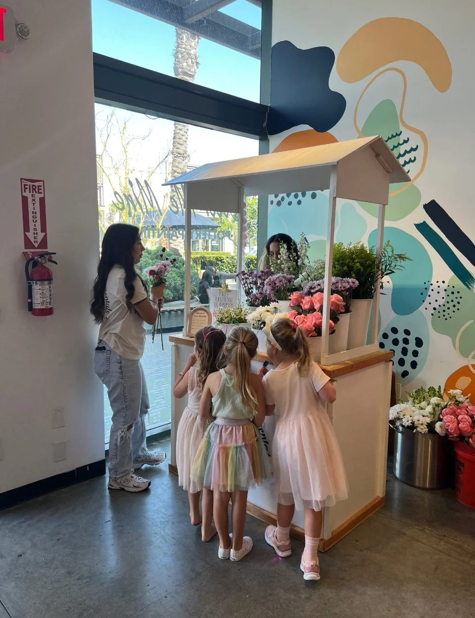 Children in pastel dresses at flower stand inside a modern store with colorful abstract wall art and large glass windows.