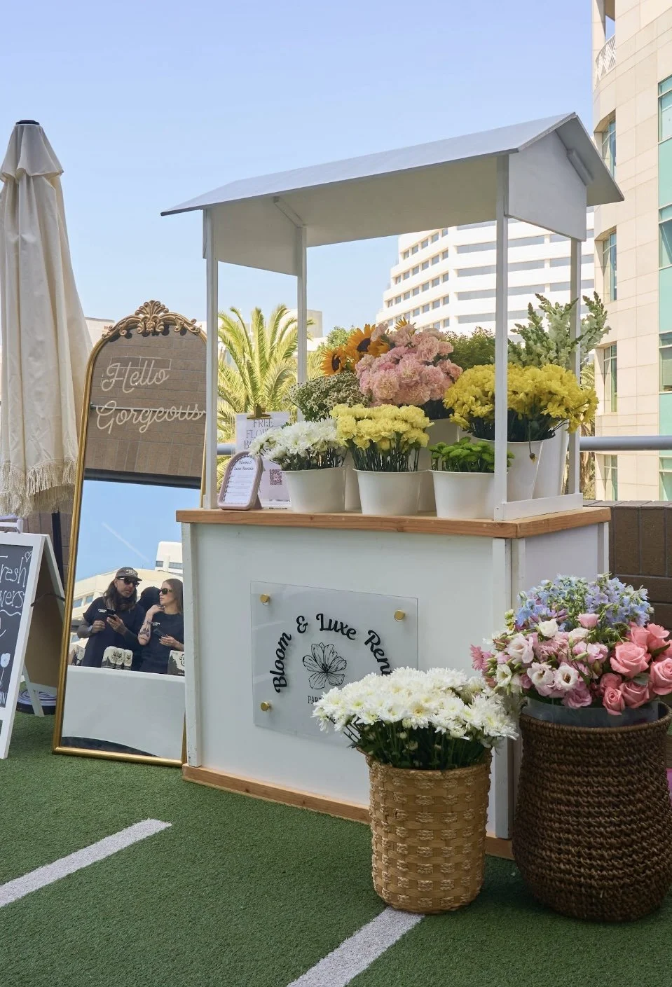 A flower stand with potted flowers on top and a sign that reads 'Bloom & Luke Review'. There are two women in the background near another stand, and a mirror with a decorative frame leaning against the flower stand that says 'Hello Gorgeous'. The scene is outdoors with high-rise buildings and palm trees behind.