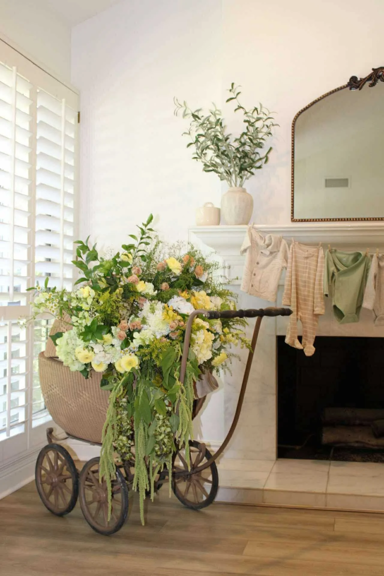 An antique wooden baby stroller filled with a large bouquet of white, yellow, and pink flowers, positioned in a bright room with a white fireplace, a mirror, and laundry hanging on a line.