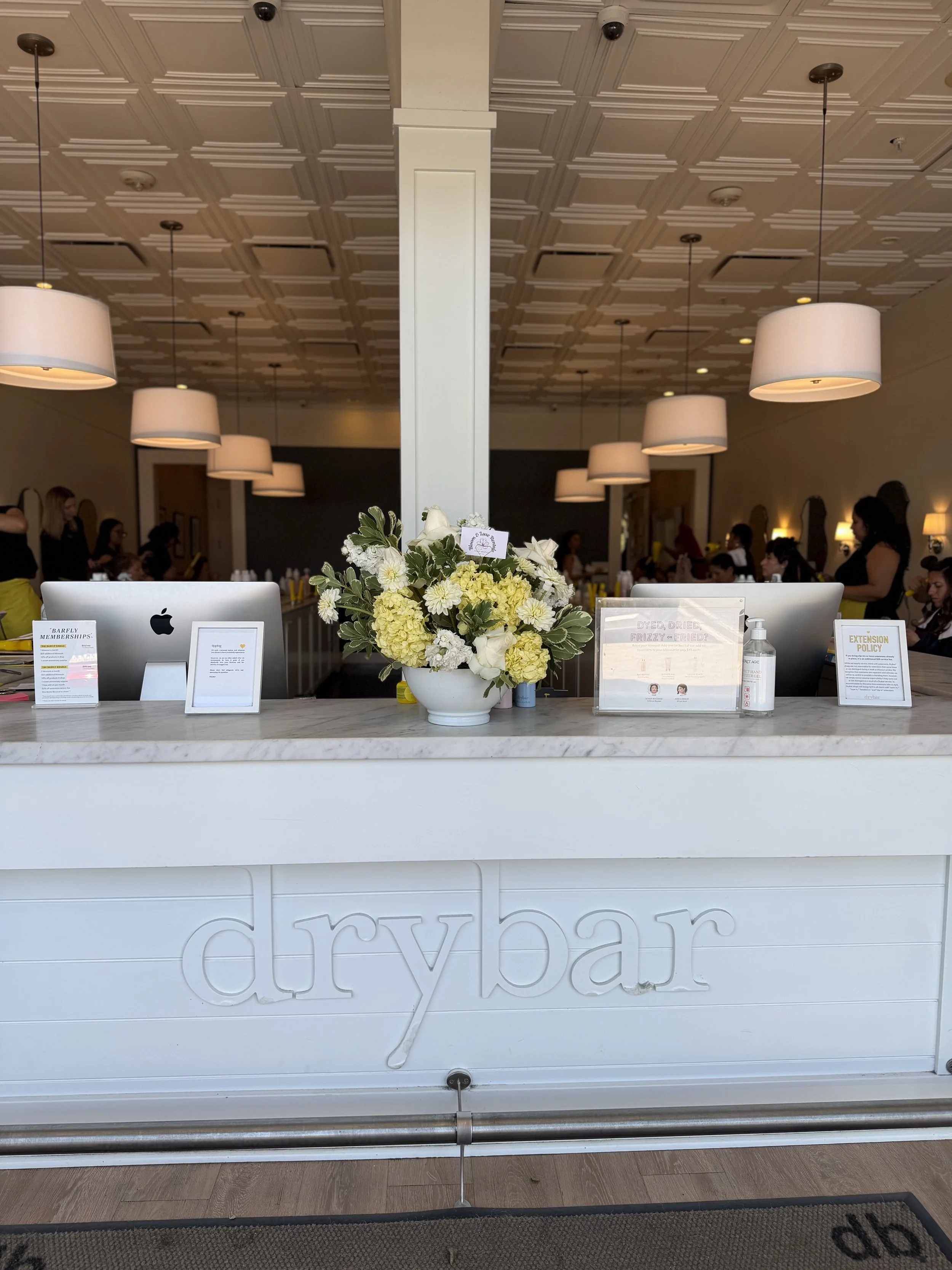 Close-up view of a bakery counter with a large white flower arrangement. The counter has the words 'dry bar' engraved on it, and various pamphlets and a bottle of hand sanitizer are displayed on top.