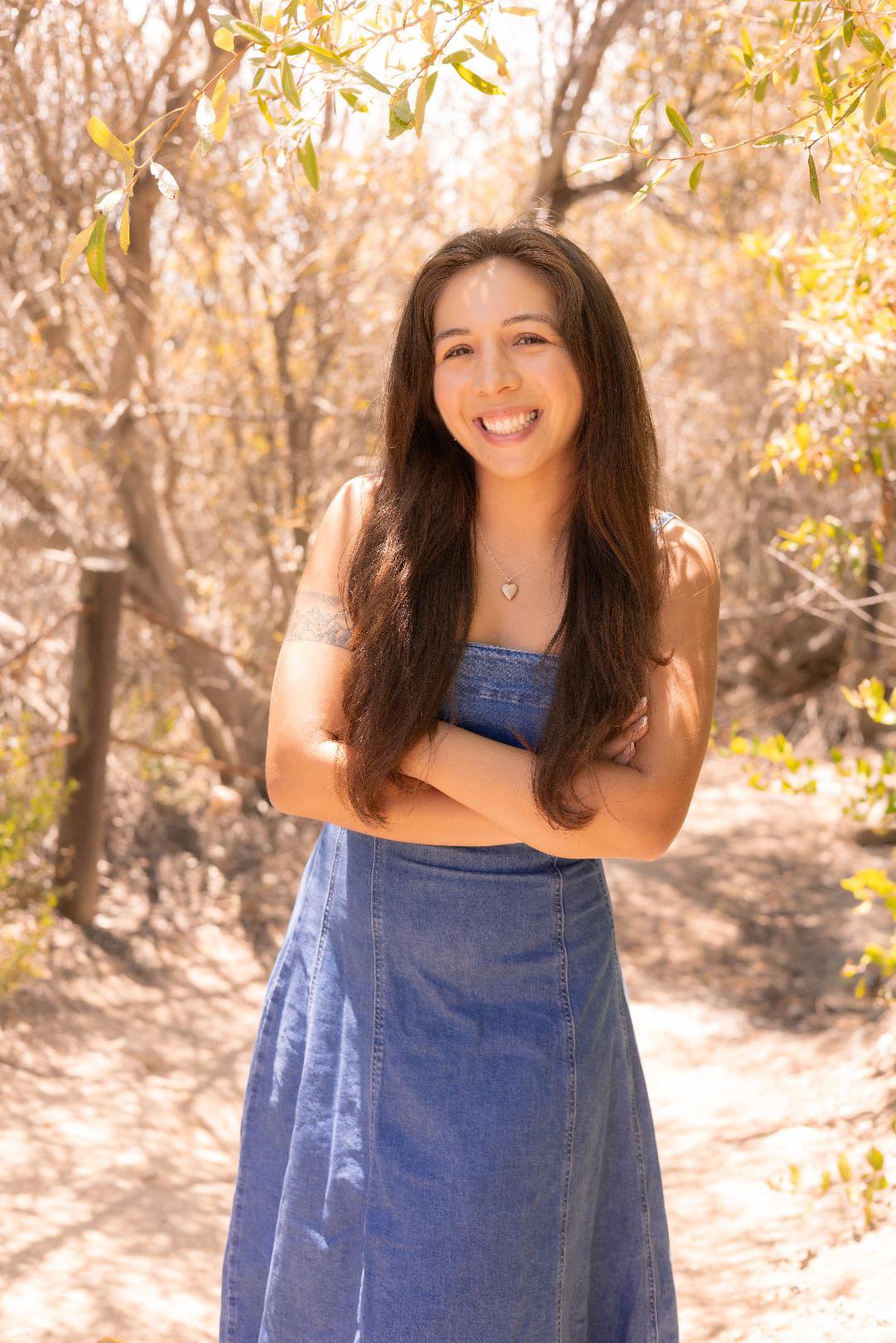 A woman with long dark hair smiling, standing with arms crossed in a sunlit outdoor setting with trees and foliage.