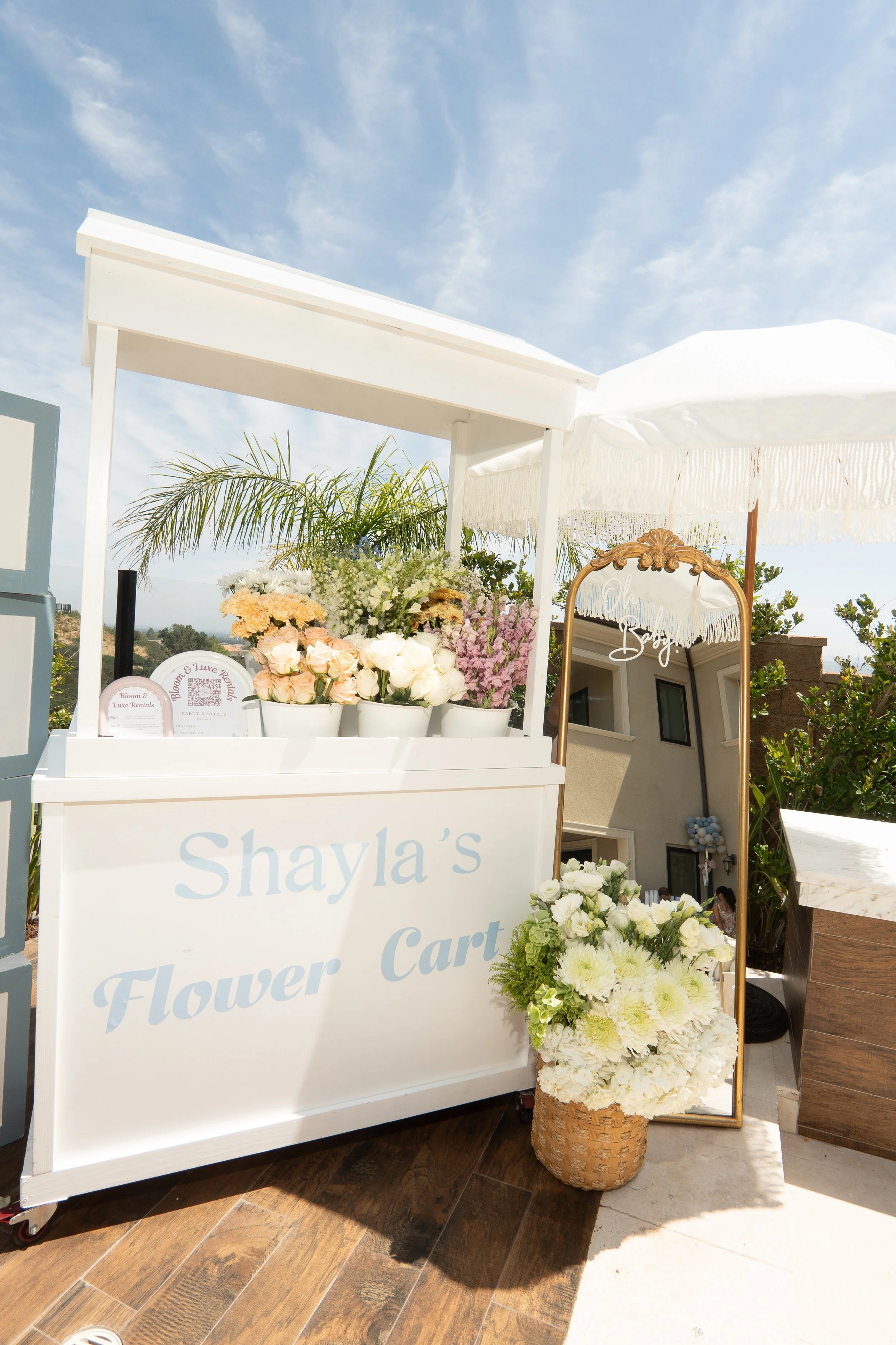 A white flower cart with the words "Shayla's Flower Cart" in light blue, decorated with various white, pink, and purple flowers, set outdoors under a blue sky with wispy clouds.