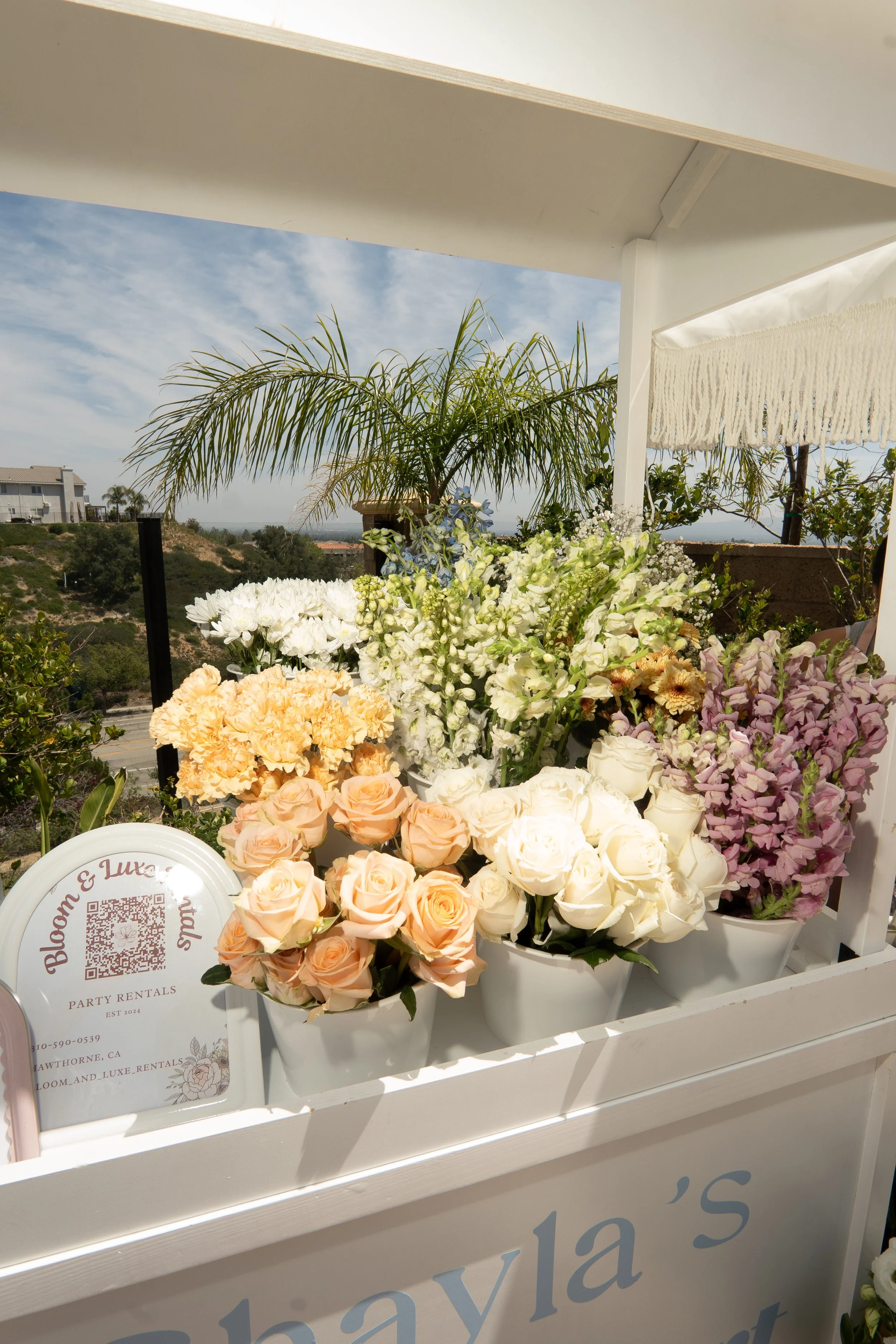 Arrangement of various pastel-colored roses, peonies, and other flowers in white buckets on a white stand, with a sign reading 'Bloom & Luxe' and a QR code, under an outdoor patio with a background view of hills, palm trees, and a partly cloudy sky.