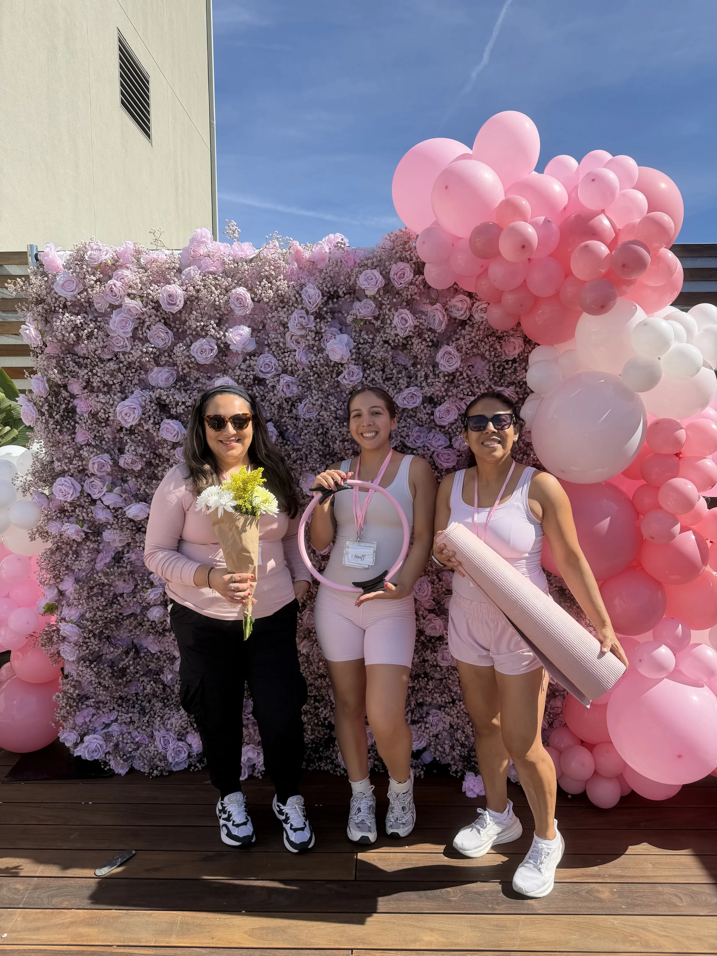 Three women standing in front of a purple floral backdrop with pink and white balloons. The woman on the left is holding a bouquet, the person in the middle is holding a pink hoop, and the woman on the right is holding a pink rolled-up mat. They are outdoors on a wooden deck with a building and blue sky in the background.