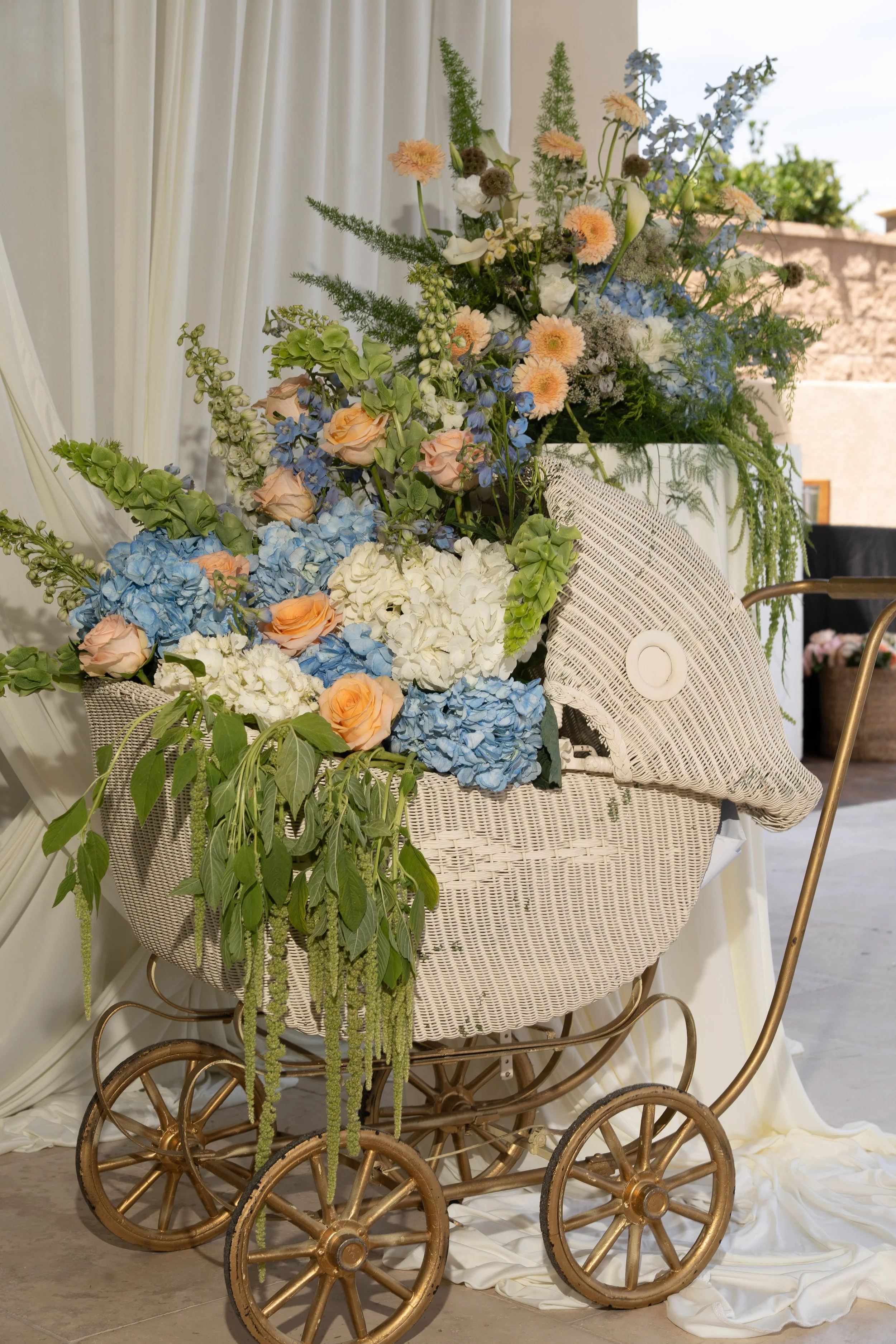 A vintage pram filled with a large arrangement of various flowers including roses, hydrangeas, and other blooms in soft pastel and blue colors, set indoors with draped white fabric and greenery.
