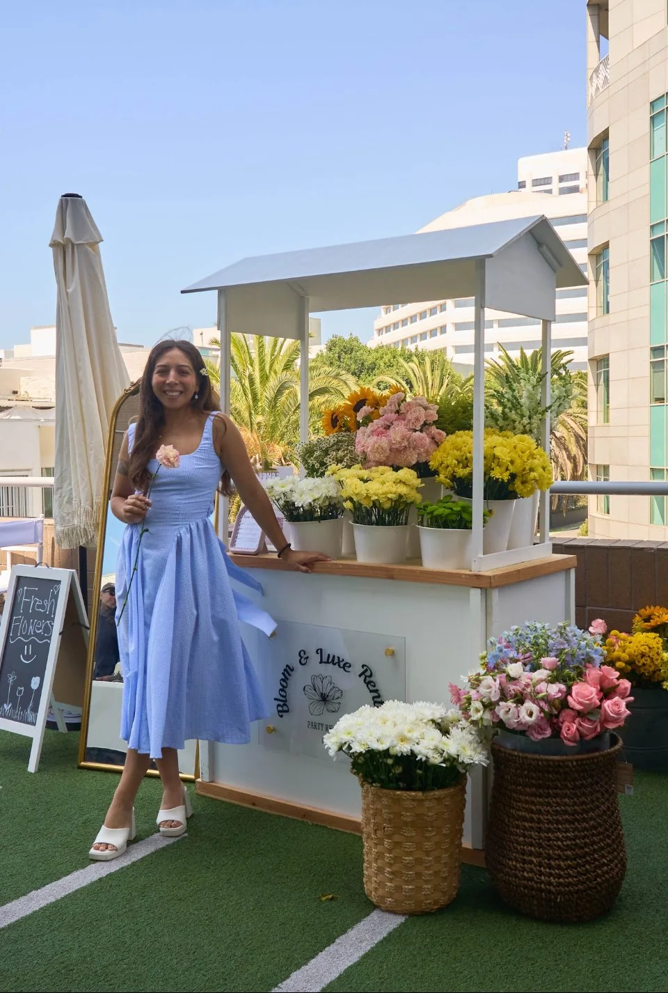 Young woman in a light blue dress standing next to a flower stand with various bright flowers and a sign that reads 'Bloom & Luxe Rez.' She is holding a pink flower and smiling. The setting appears to be an outdoor event with buildings and palm trees