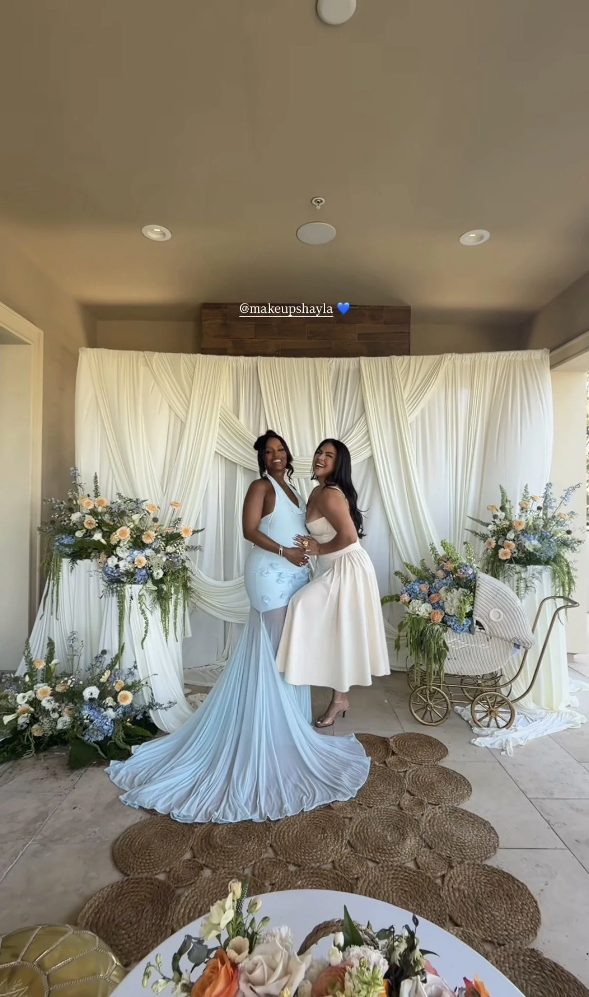Two women in elegant dresses posing in front of a decorated backdrop with flowers, draped fabric, and greenery, at what appears to be a celebration or wedding event.
