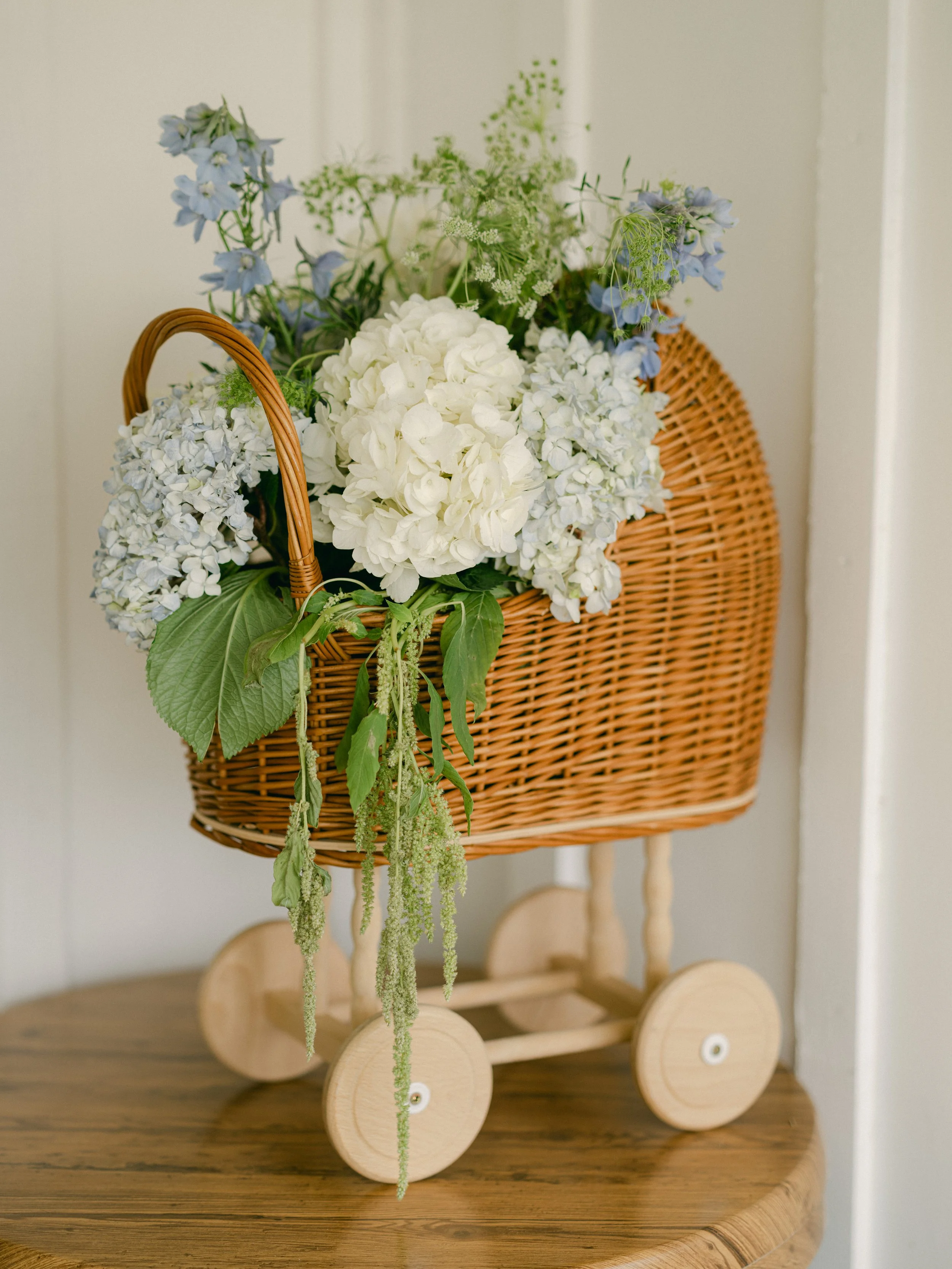 A decorative baby's doll stroller filled with white and blue flowers and greenery, placed on a wooden table.