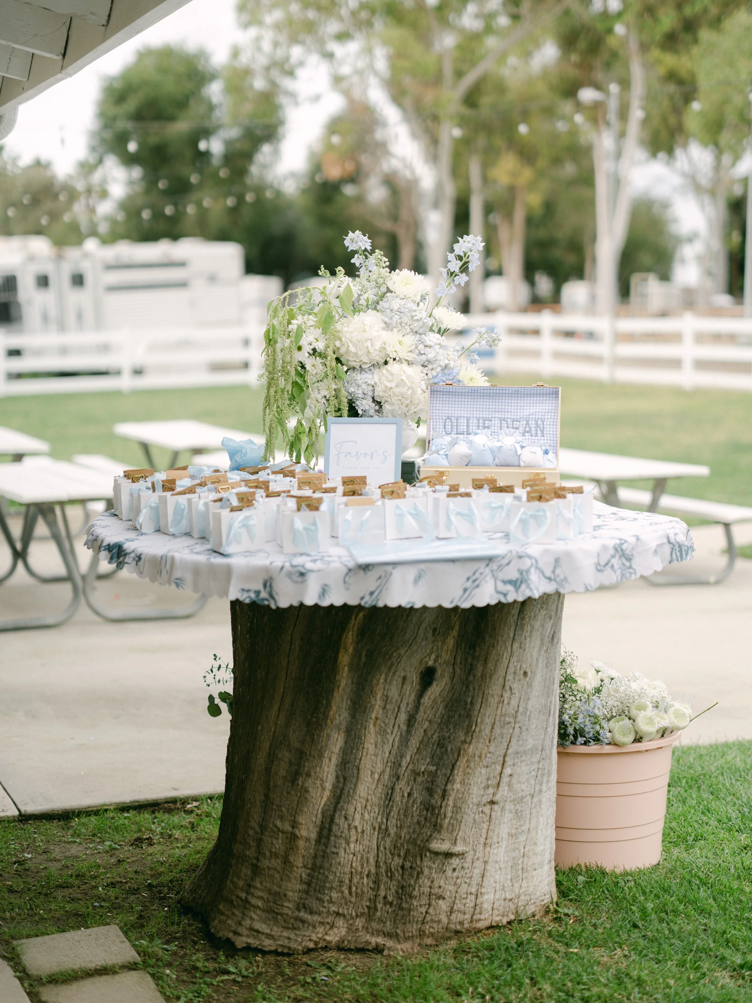 A table with white flowers and small gift boxes outdoors, next to a large tree with a trunk as a table base, in a garden setting.