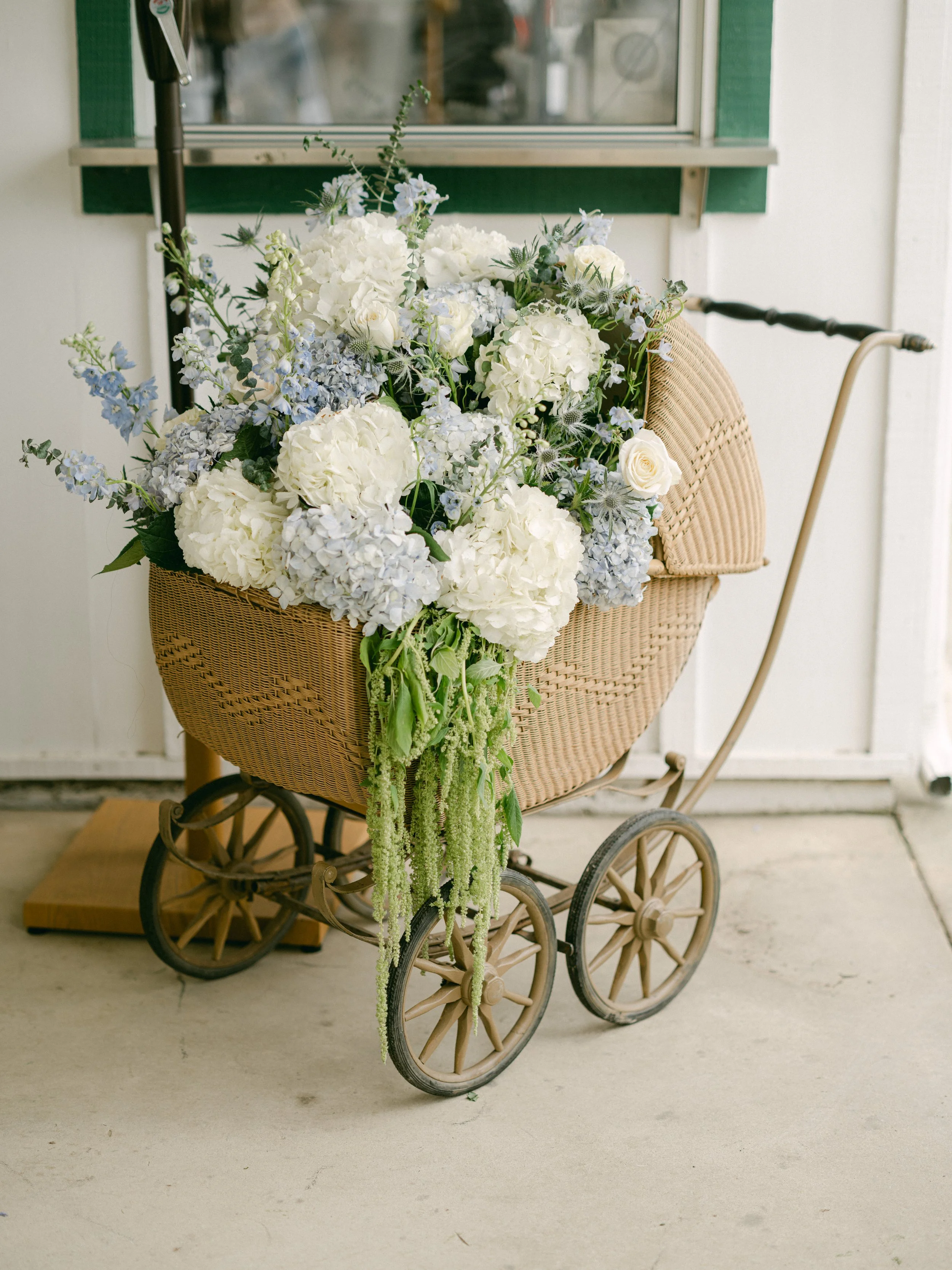 A vintage baby stroller filled with white and light purple flowers, including hydrangeas, roses, and other greenery, positioned on a concrete surface near a window with green trim.