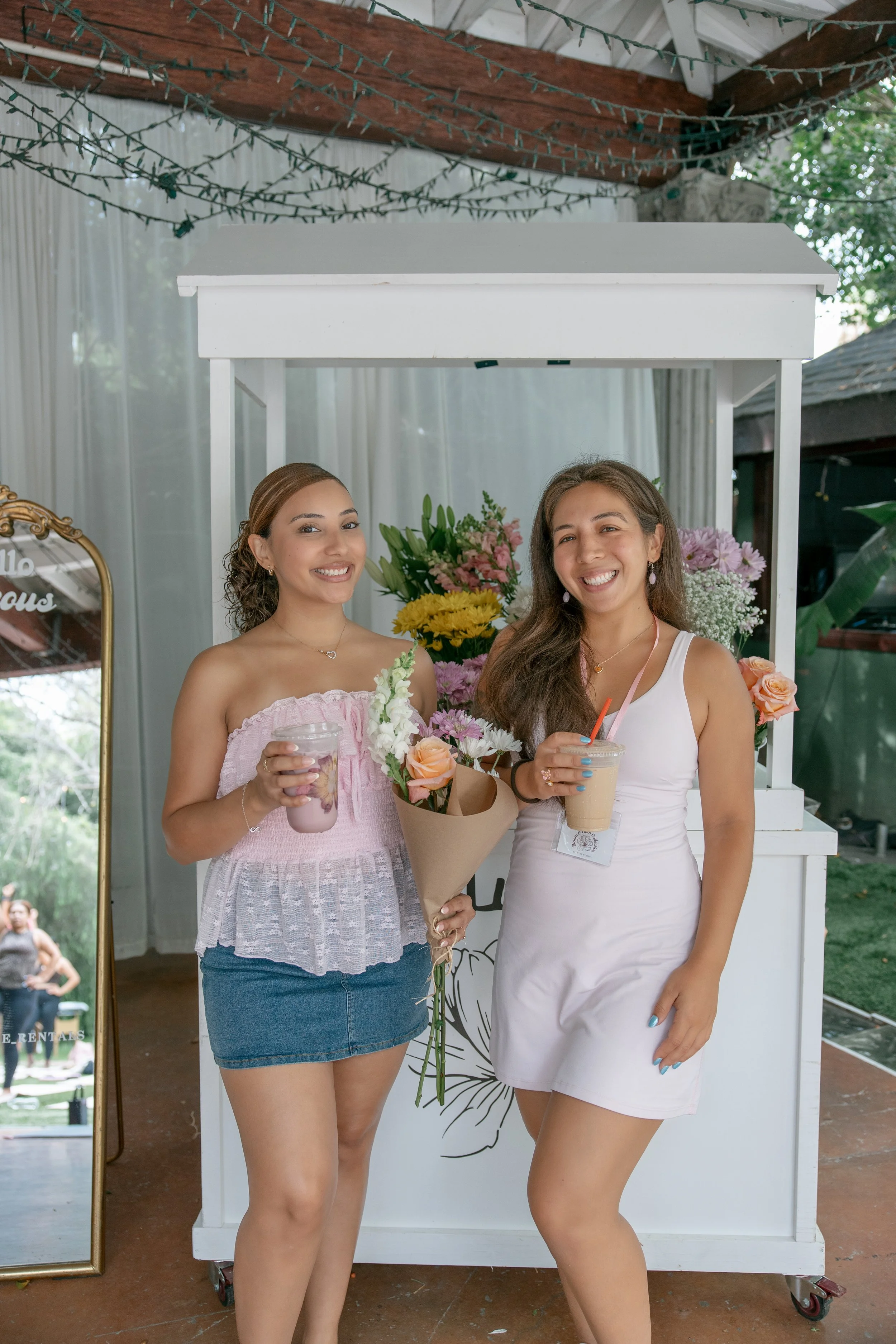 Two women standing in front of a white floral photo booth, holding drinks and smiling at the camera. One woman is holding a bouquet of flowers.