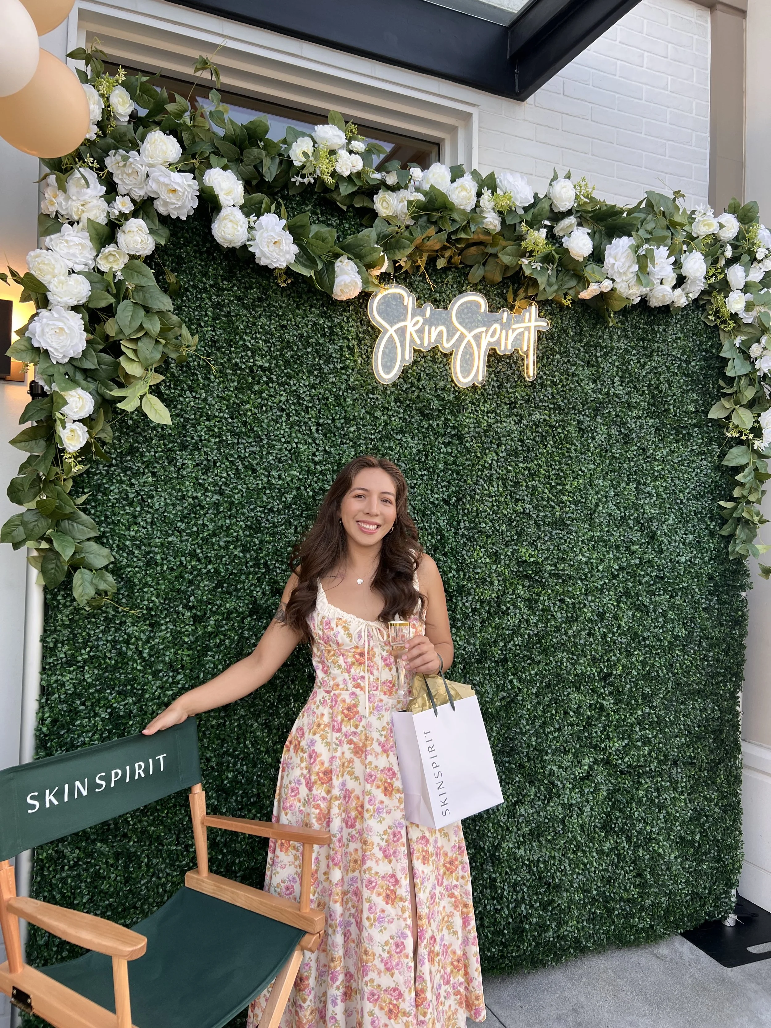 A woman with long, wavy brown hair, wearing a floral dress and holding a glass of champagne, standing in front of a lush green hedge wall with white flowers and a illuminated sign that reads 'SkinSpirit'.