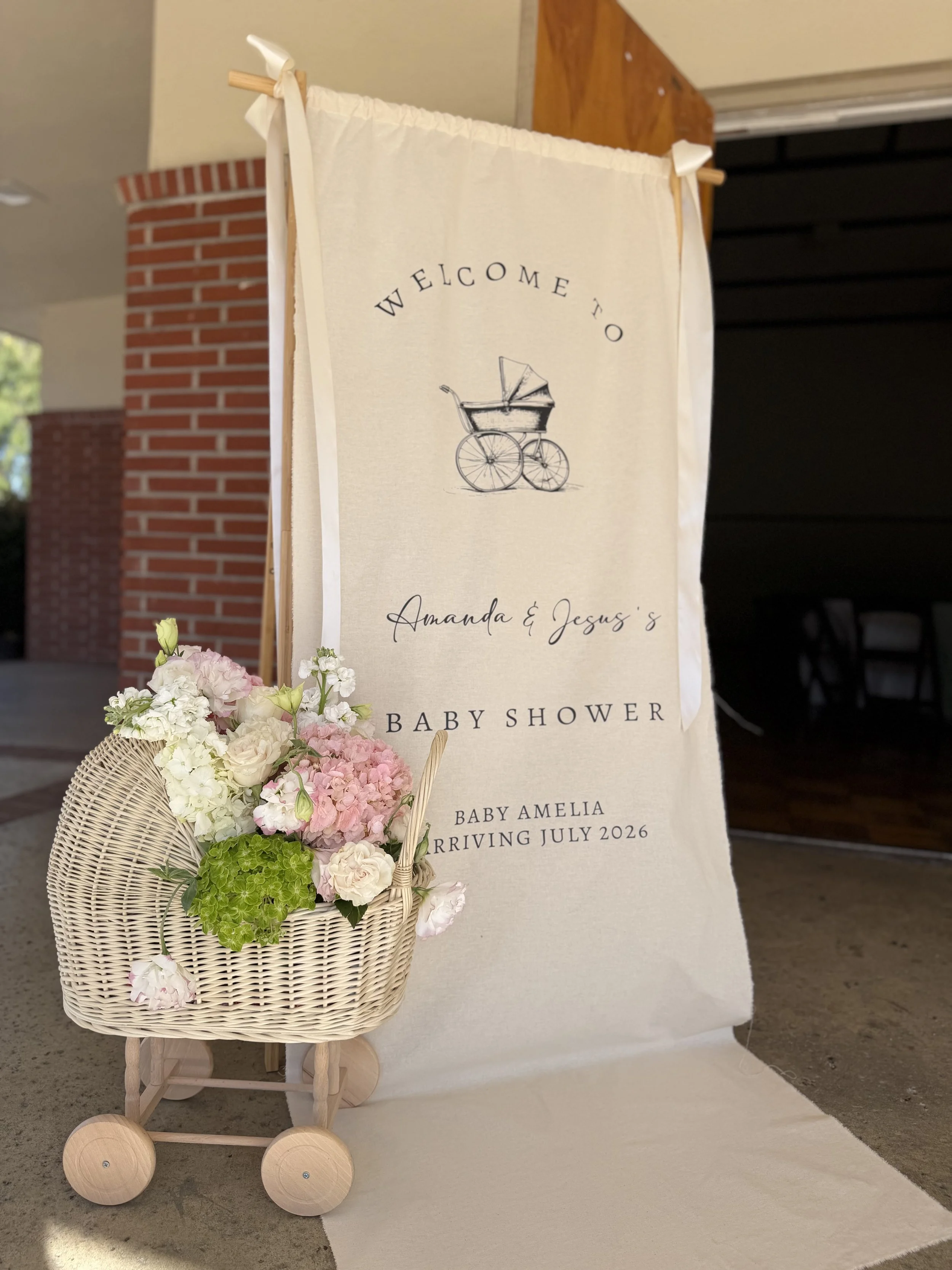 A baby shower display with a cream-colored banner and a small wicker stroller filled with pink, white, and green flowers, and a welcome message for baby Amelia arriving in July 2026.