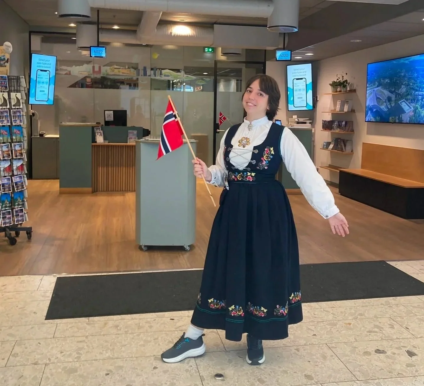 Woman dressed in traditional Norwegian clothing holding a small Norwegian flag inside a building, possibly an airport or a public reception area.