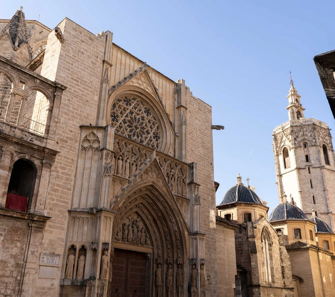 Facade of a historic Gothic-style cathedral with intricate stone carvings and large stained glass windows, a tall bell tower, and smaller domed structures against a clear blue sky.