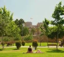 A park with green grass, trees, and people relaxing on a blanket. In the background, there is a large castle-like building.