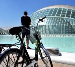 A man with a bicycle looking at a modern glass building with a curved design, near a water feature, on a sunny day.