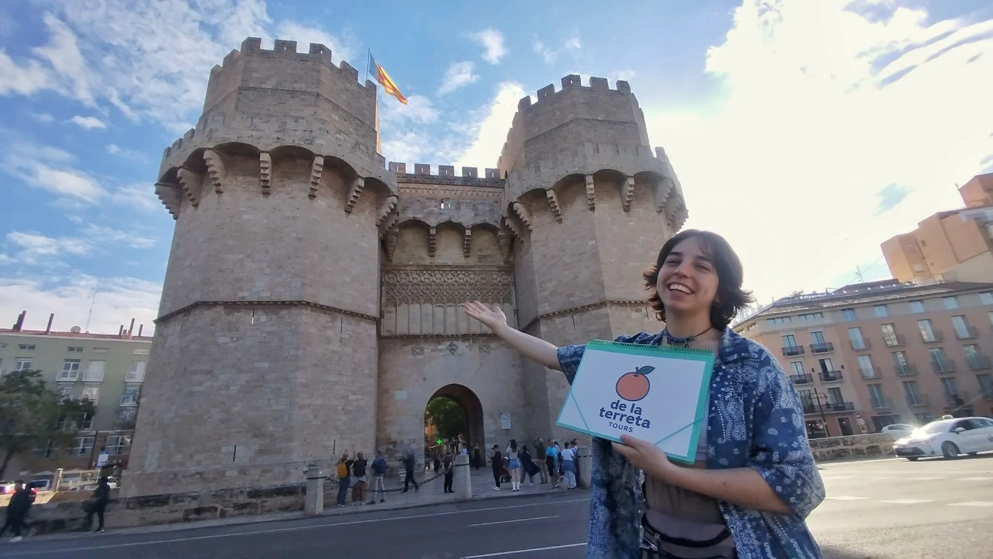 A person smiling and gesturing towards a historical stone gate with two towers, a flag flying atop, in an urban setting during daytime.