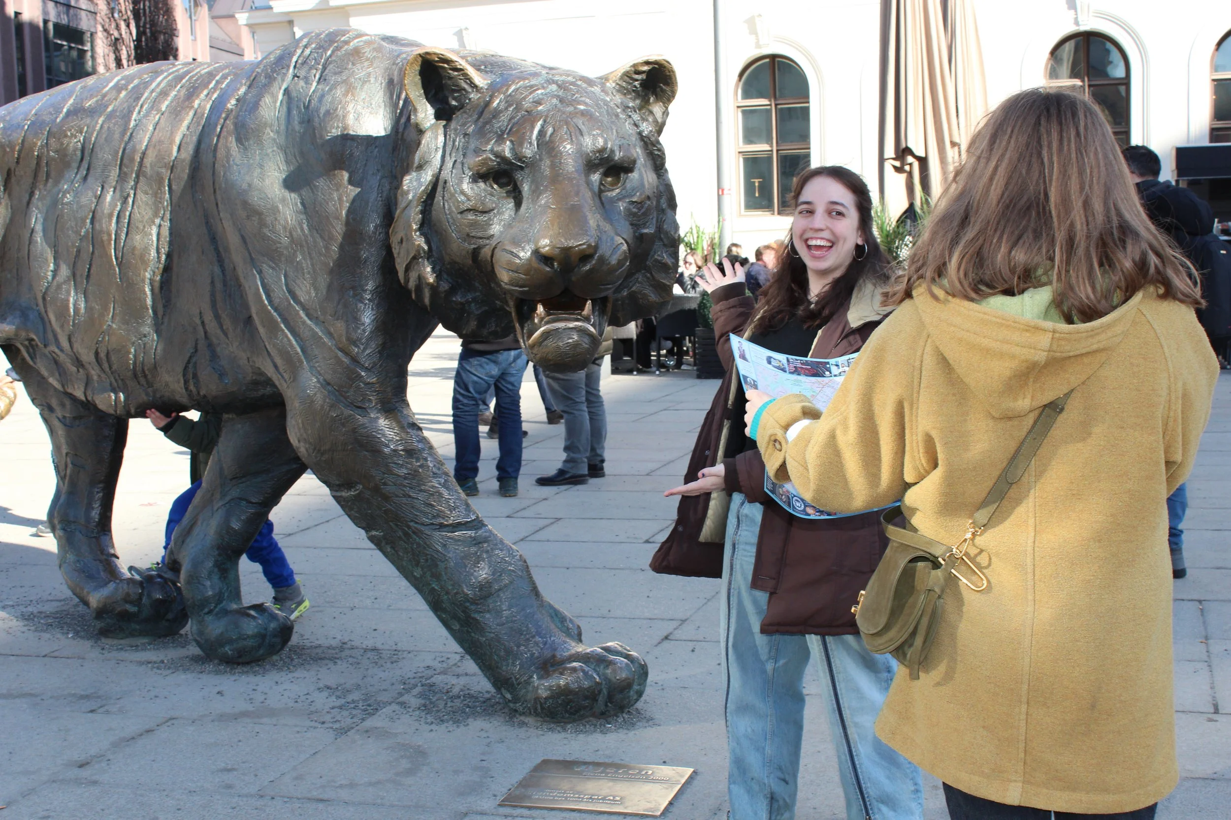 A bronze statue of a lion in a walking stance with its mouth slightly open. A woman with curly hair, hoop earrings, and a dark jacket is smiling and looking at the camera, standing near the lion statue. Another woman with straight hair, wearing a yellow coat, is facing the statue with her back to the camera. There are other people in the background, and the scene appears to be in an outdoor urban setting with buildings and windows.