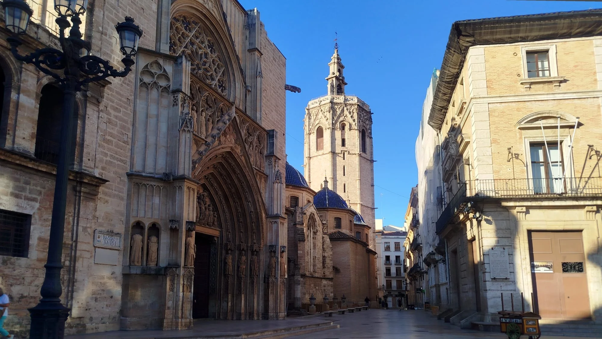 Street view showing historic buildings with detailed architecture, including Gothic and baroque elements, and a tall church tower topped with a small spire under a clear blue sky.