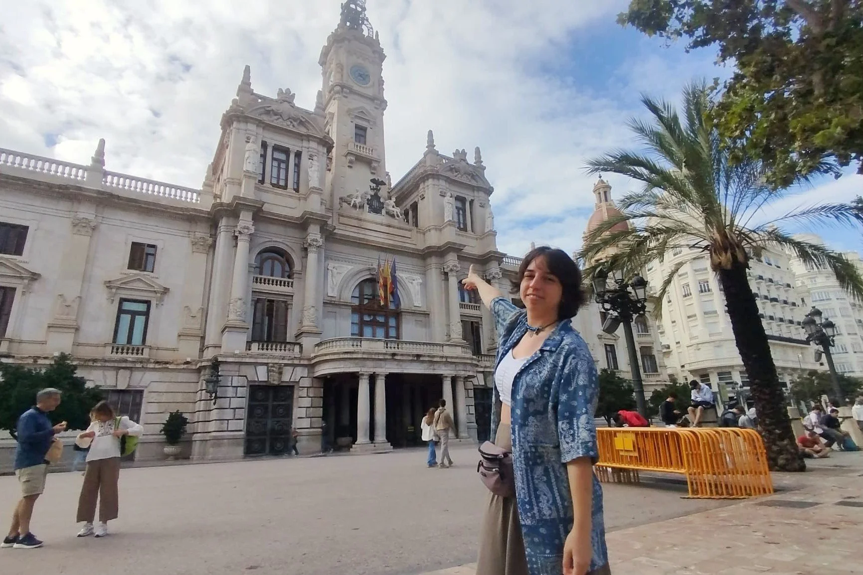 A woman pointing at a historic building with a clock tower in a city square. The building has a neoclassical architectural style with flags hanging on it. There are people walking and sitting in the square, with a palm tree on the right side of the image.