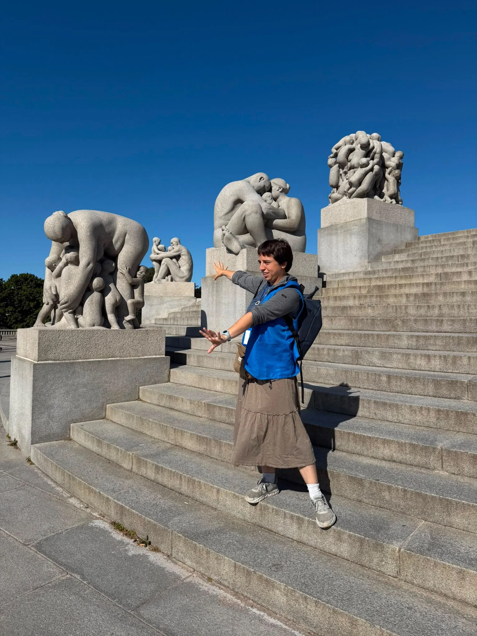 A man in a blue vest and a long brown skirt poses on stone steps in front of large sculptures depicting human figures engaged in various activities. The sculptures are outdoors against a clear blue sky.
