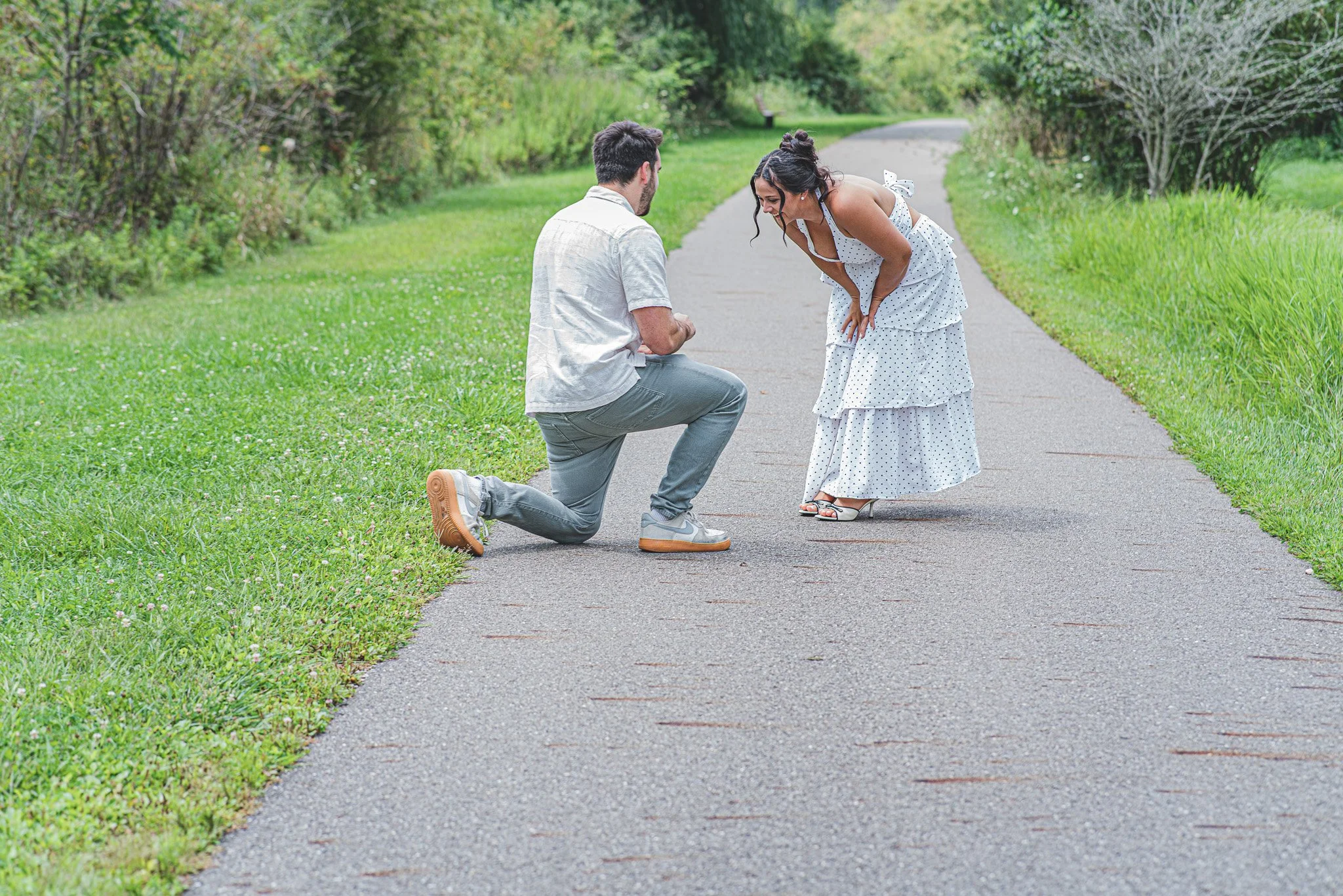 A man proposes marriage to a woman on a park pathway. The man is kneeling on one knee, holding a ring, while the woman bends forward in surprise. The setting is outdoors with green grass and trees in the background.