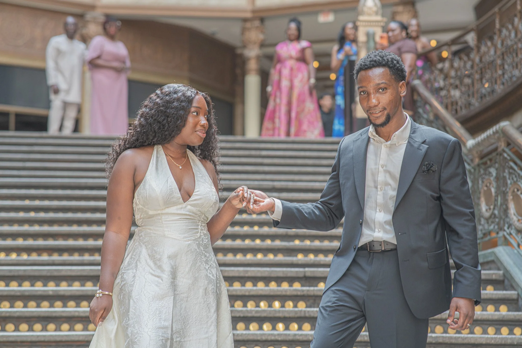 A couple dressed in formal attire, holding hands and smiling, standing on the stairs inside a grand building, possibly a wedding or special event, with several women in colorful dresses in the background.