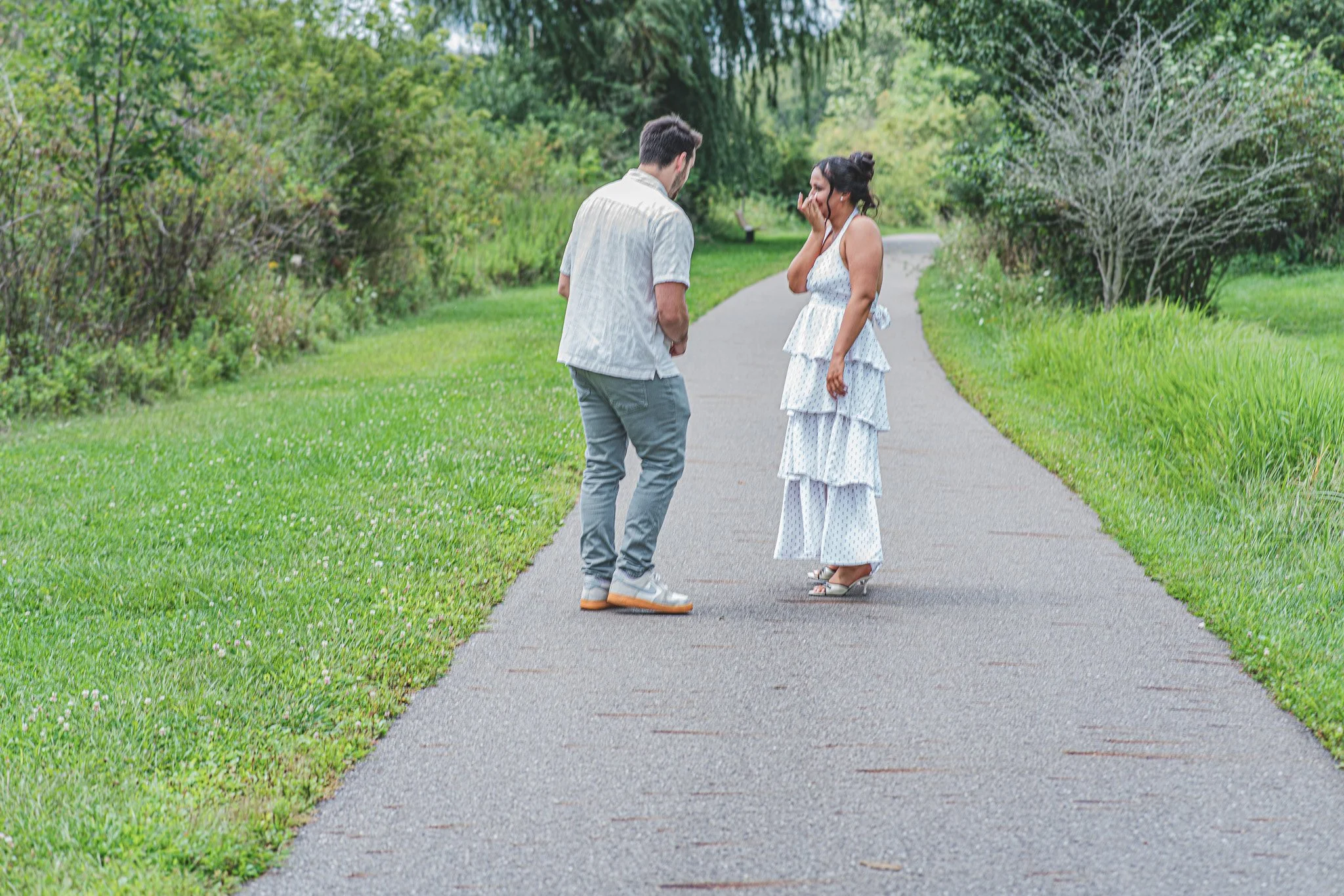 A man and a woman stand on a park pathway, engaged in a conversation, surrounded by green grass and trees.