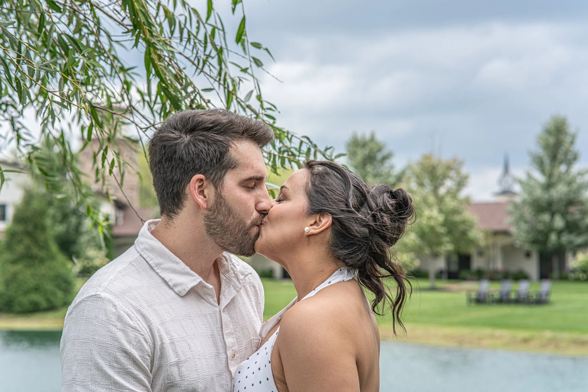 A couple is kissing outdoors near a lake, with trees and a building in the background under a cloudy sky.