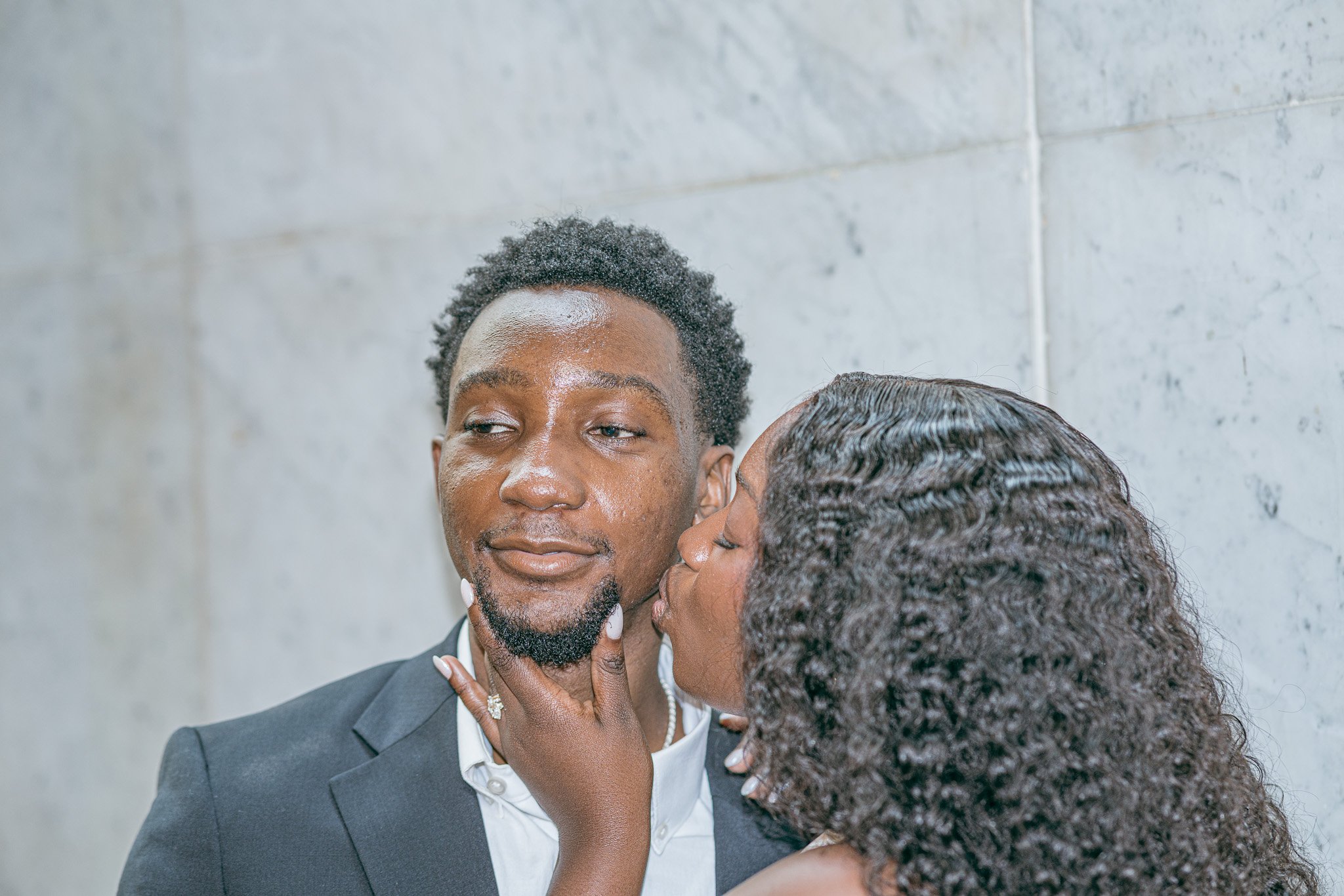 A man in a suit, with short curly hair and a beard, is receiving a kiss on the cheek from a woman with long curly hair. The background is a plain gray wall.