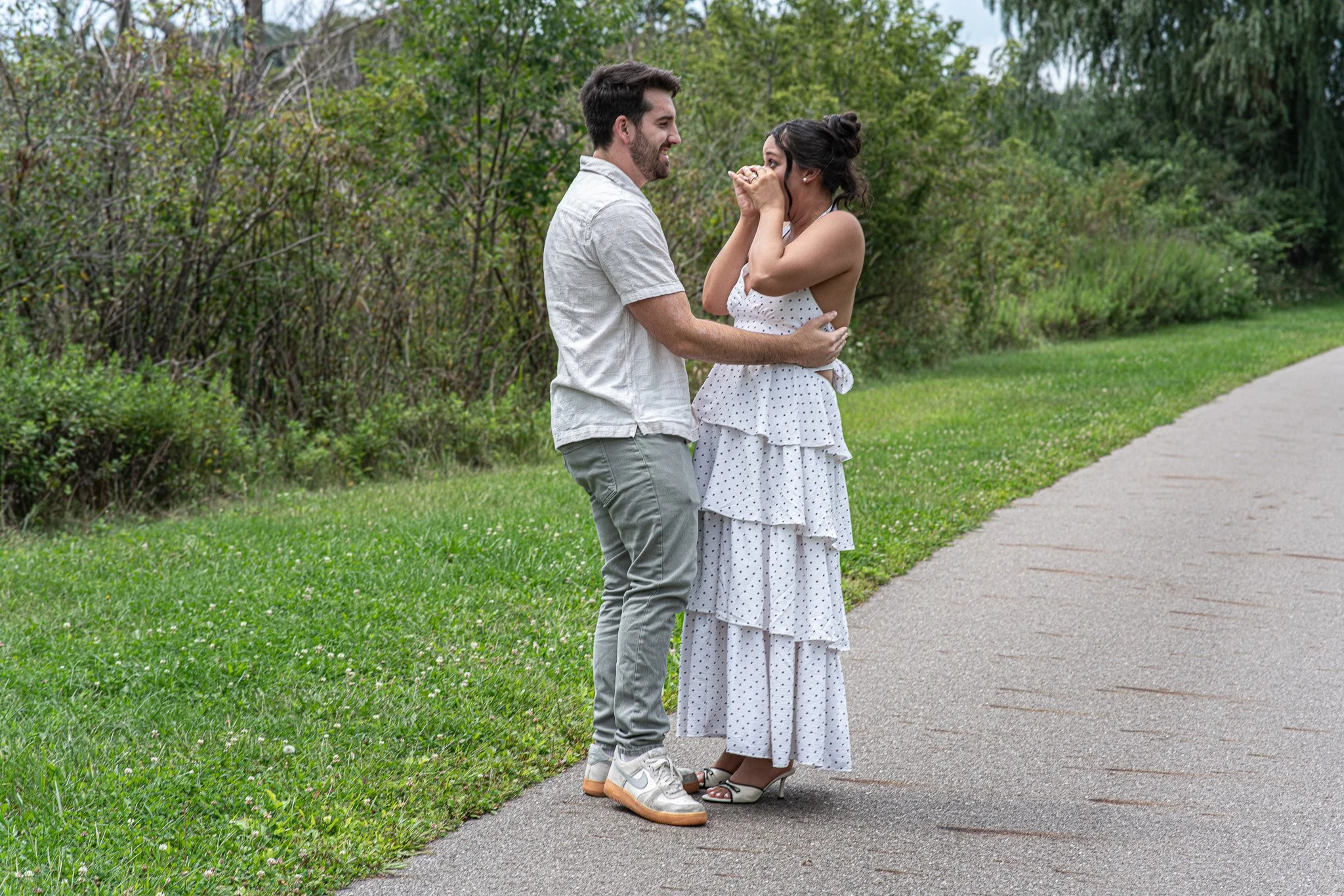 A man and woman standing close together outdoors on a path, smiling and about to kiss, with greenery in the background.