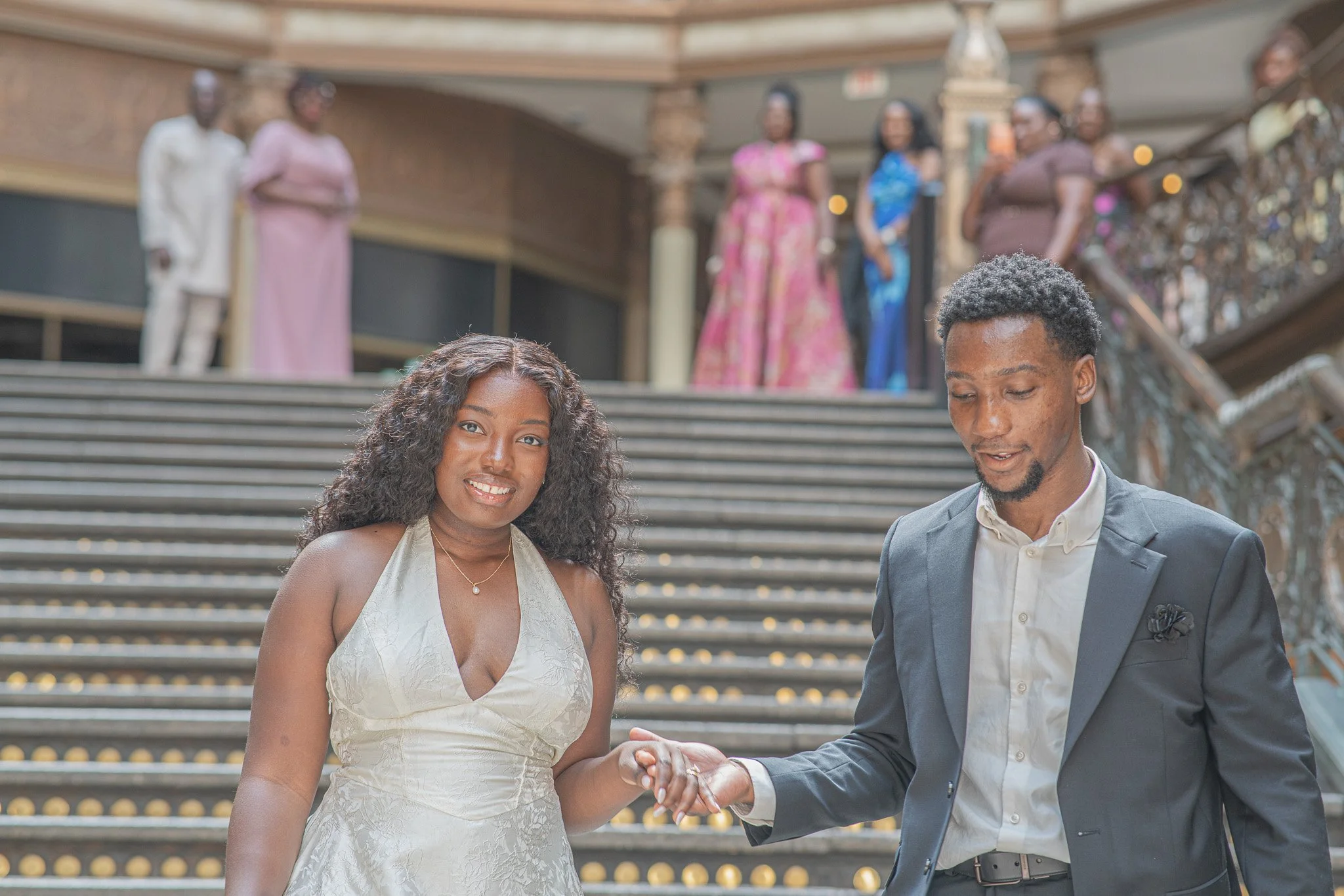 A couple dressed in wedding attire standing on steps, holding hands, with blurred women in dresses standing in the background.