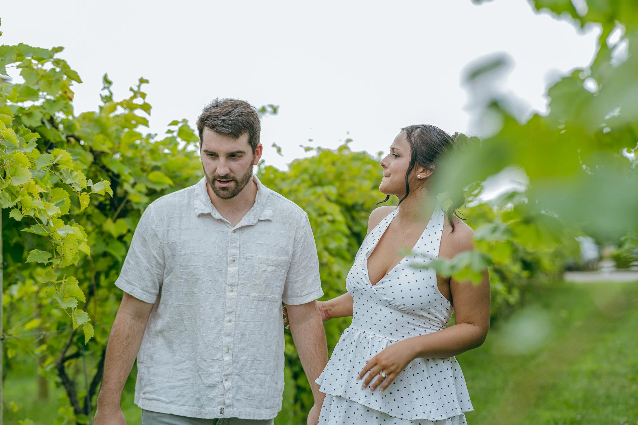 A man and woman walking through a lush green vineyard. The man has short brown hair, a beard, and wears a light-colored shirt. The woman has dark hair styled in waves, wearing a white polka dot dress. They appear to be engaged in conversation.