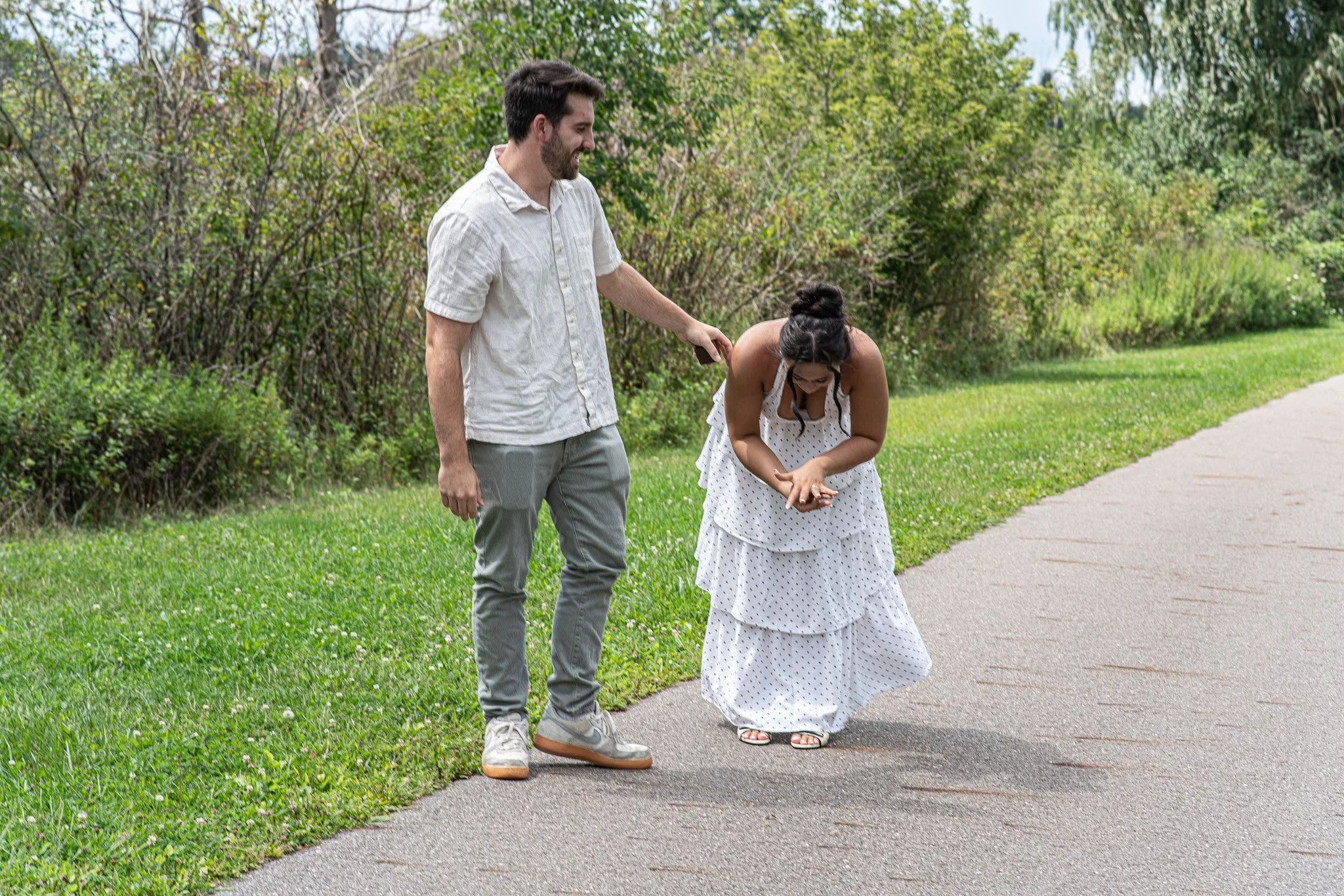 A man in a light-colored shirt and gray pants is smiling and holding a woman's hand as she bends over, wearing a white polka-dot dress, on a sidewalk with greenery in the background.