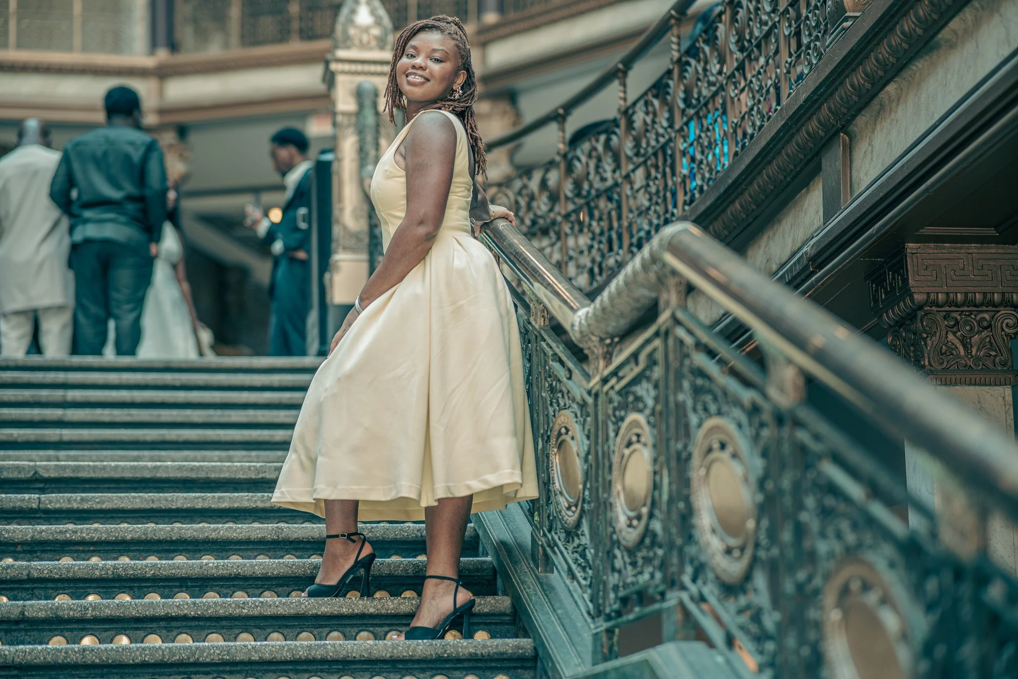 Woman in a light yellow dress and black high heels standing on ornate staircase railing with others in formal attire in background.