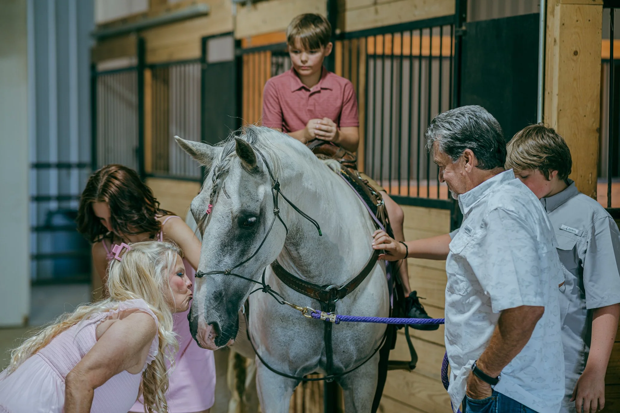 Group of children and adults gathered around the families gray horse in a stable.
