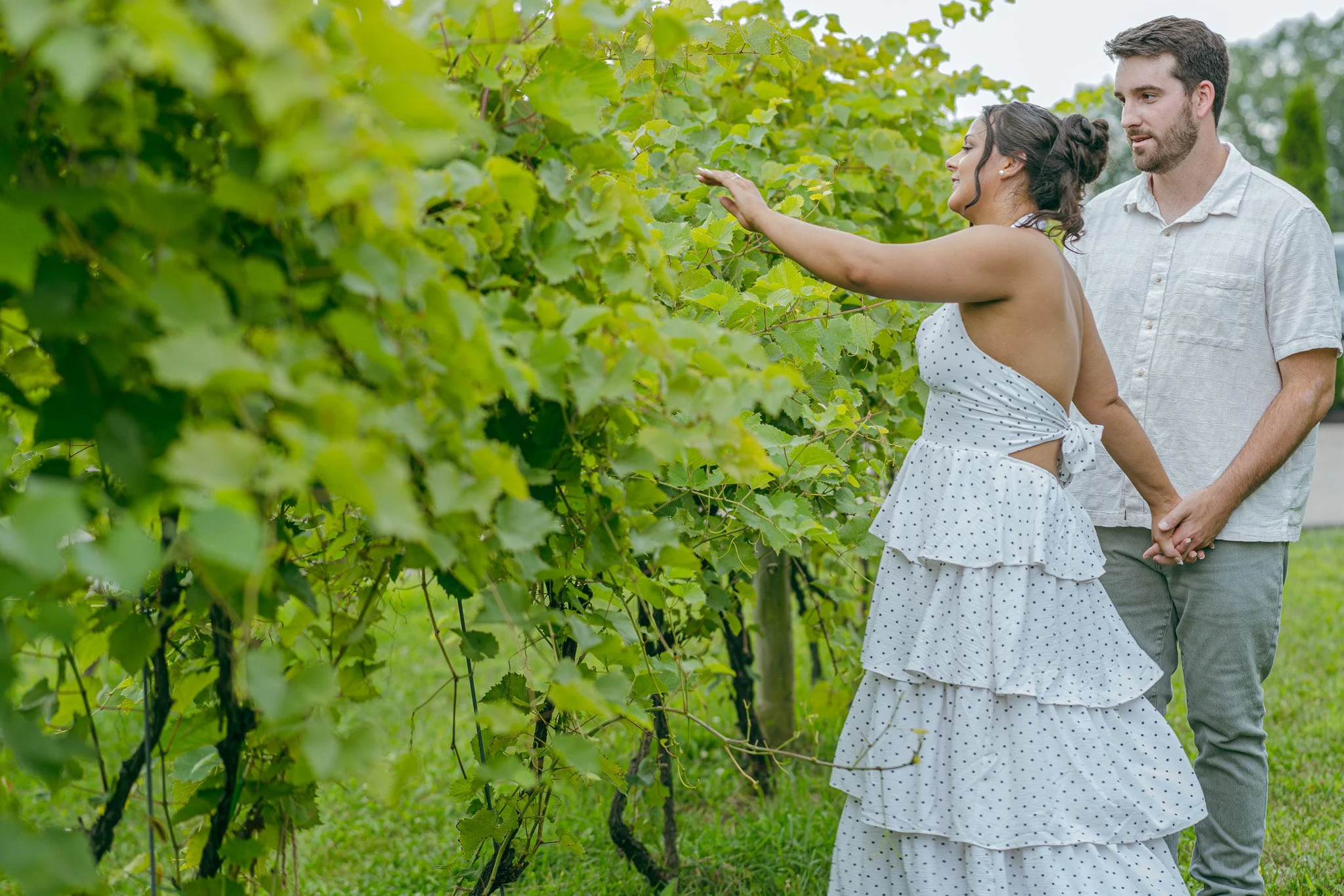 A woman in a white polka dot dress reaching out to touch grapevines, with a man standing behind her in a vineyard.
