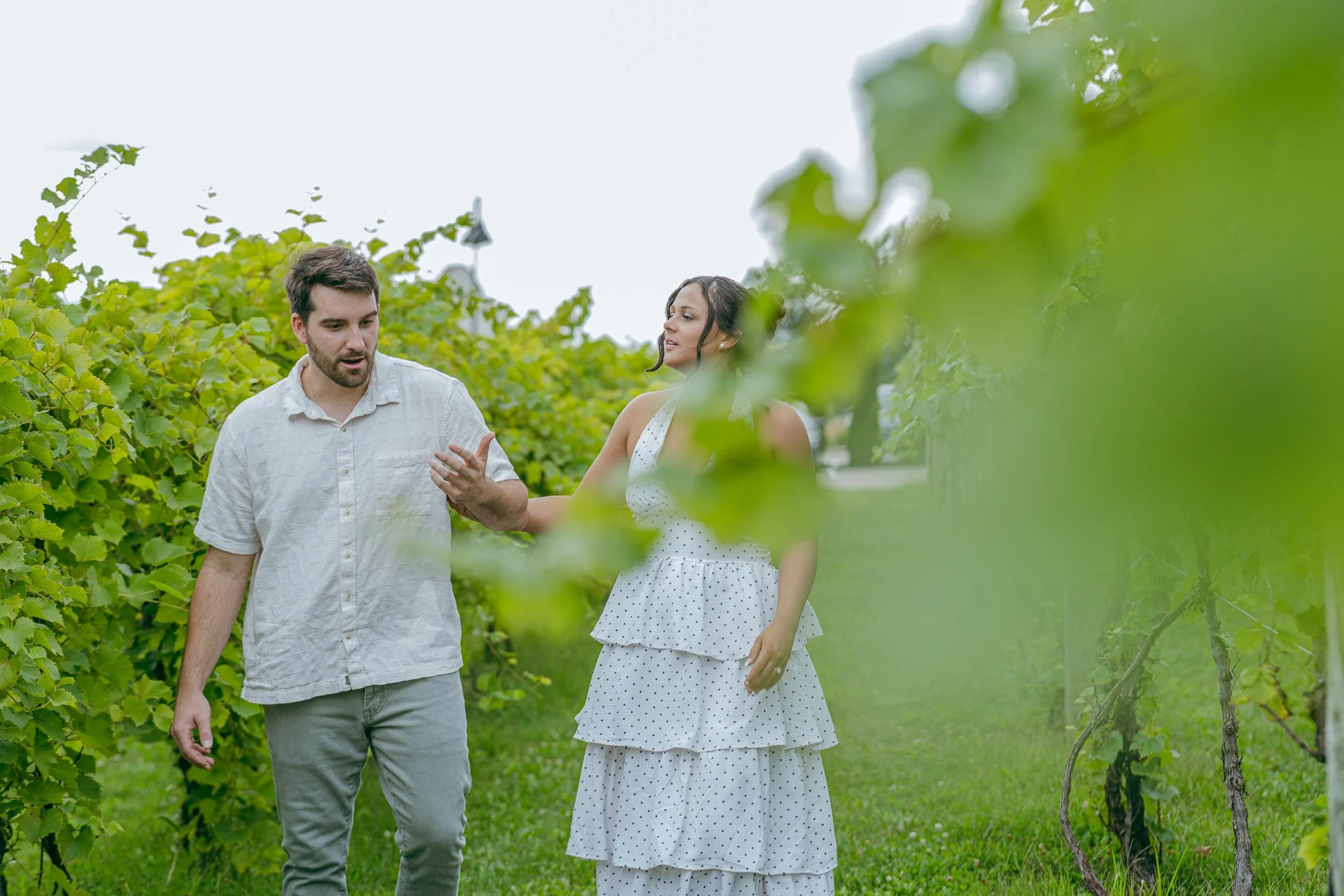 A man and a woman walking through a lush green vineyard, engaging in conversation, surrounded by grapevines and foliage.